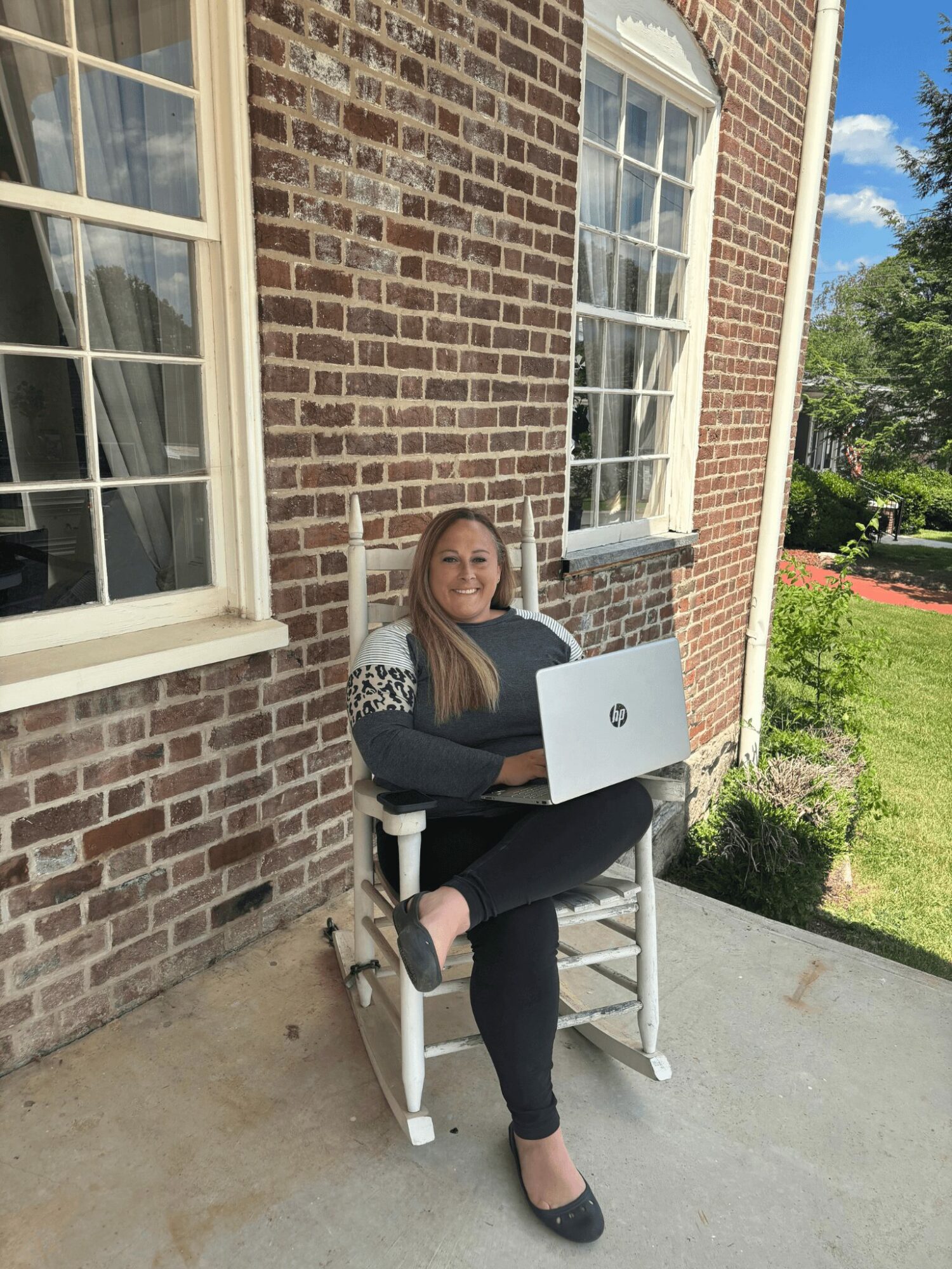 Woman sitting on a white chair outside a brick building with large windows, using a laptop, with trees and blue sky in background.