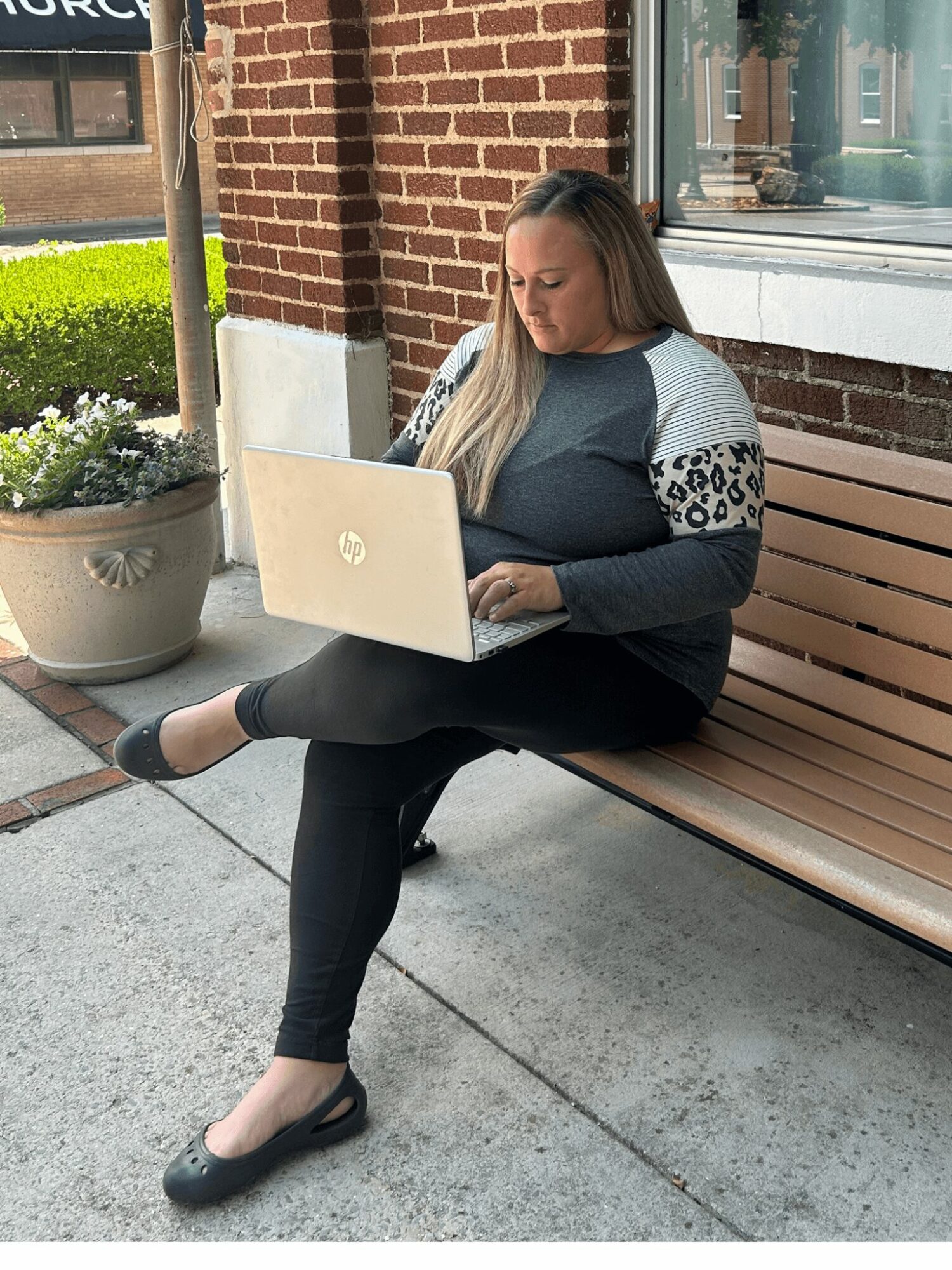 Woman sitting on a bench outside, working on a laptop, with a brick building and potted plants nearby.