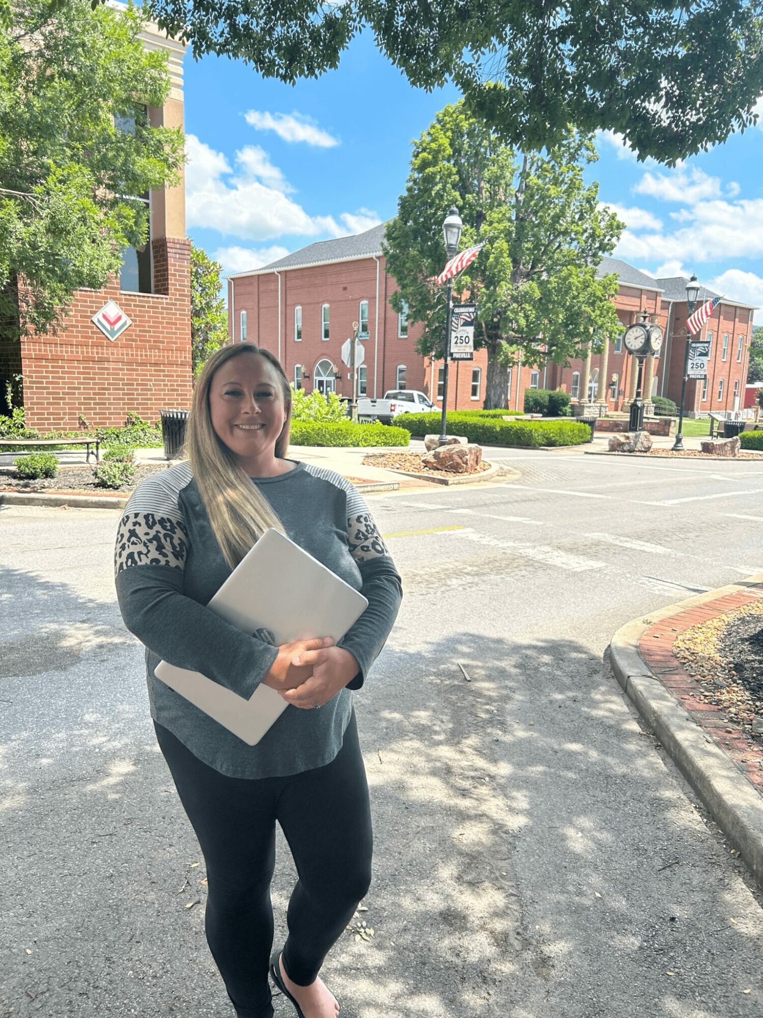 Woman standing outdoors holding a folder, trees and brick buildings in the background, sunny day with blue sky.
