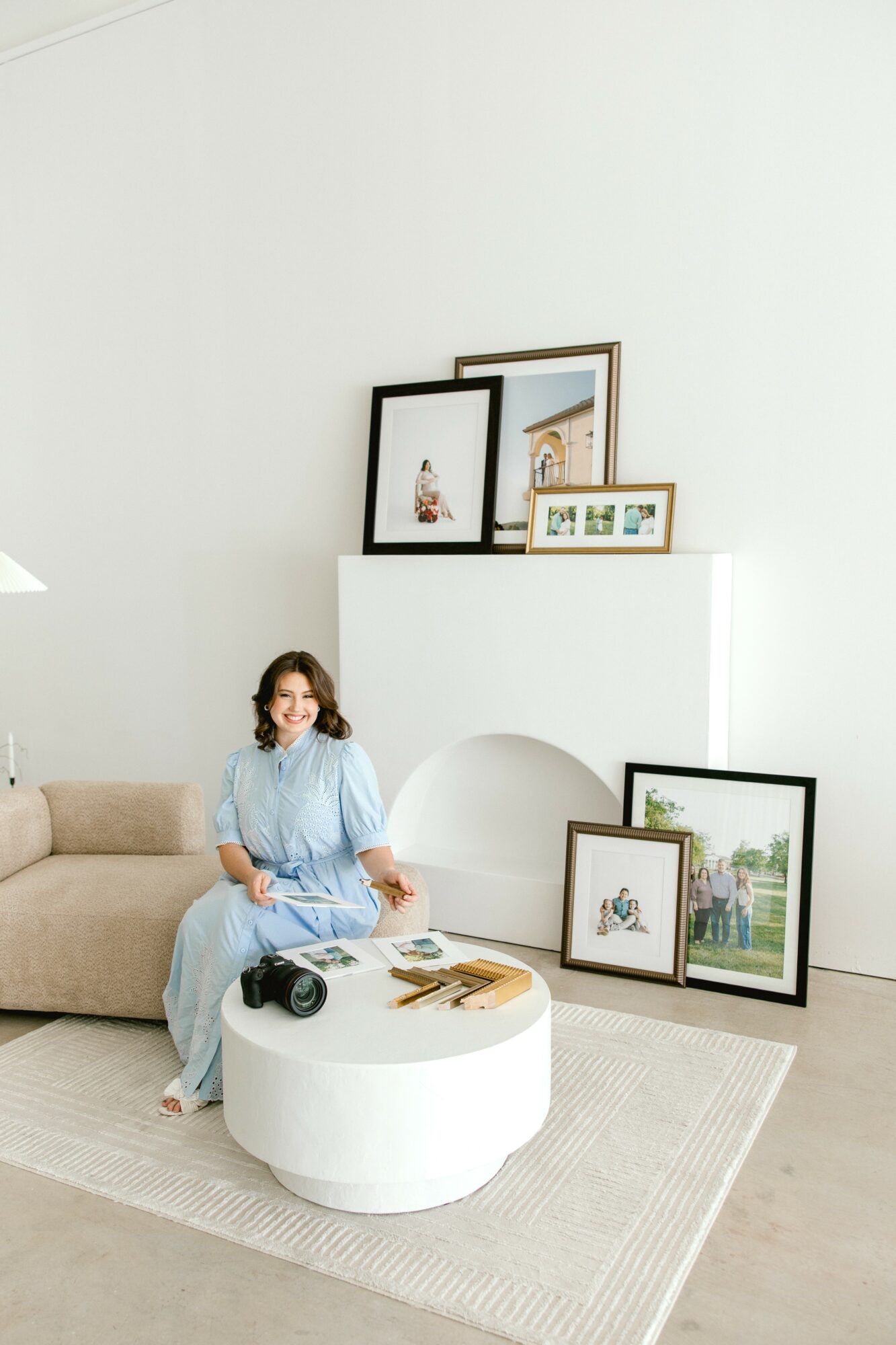 Woman sitting on a beige sofa in a bright room with framed pictures on the wall and floor, and a white round coffee table.