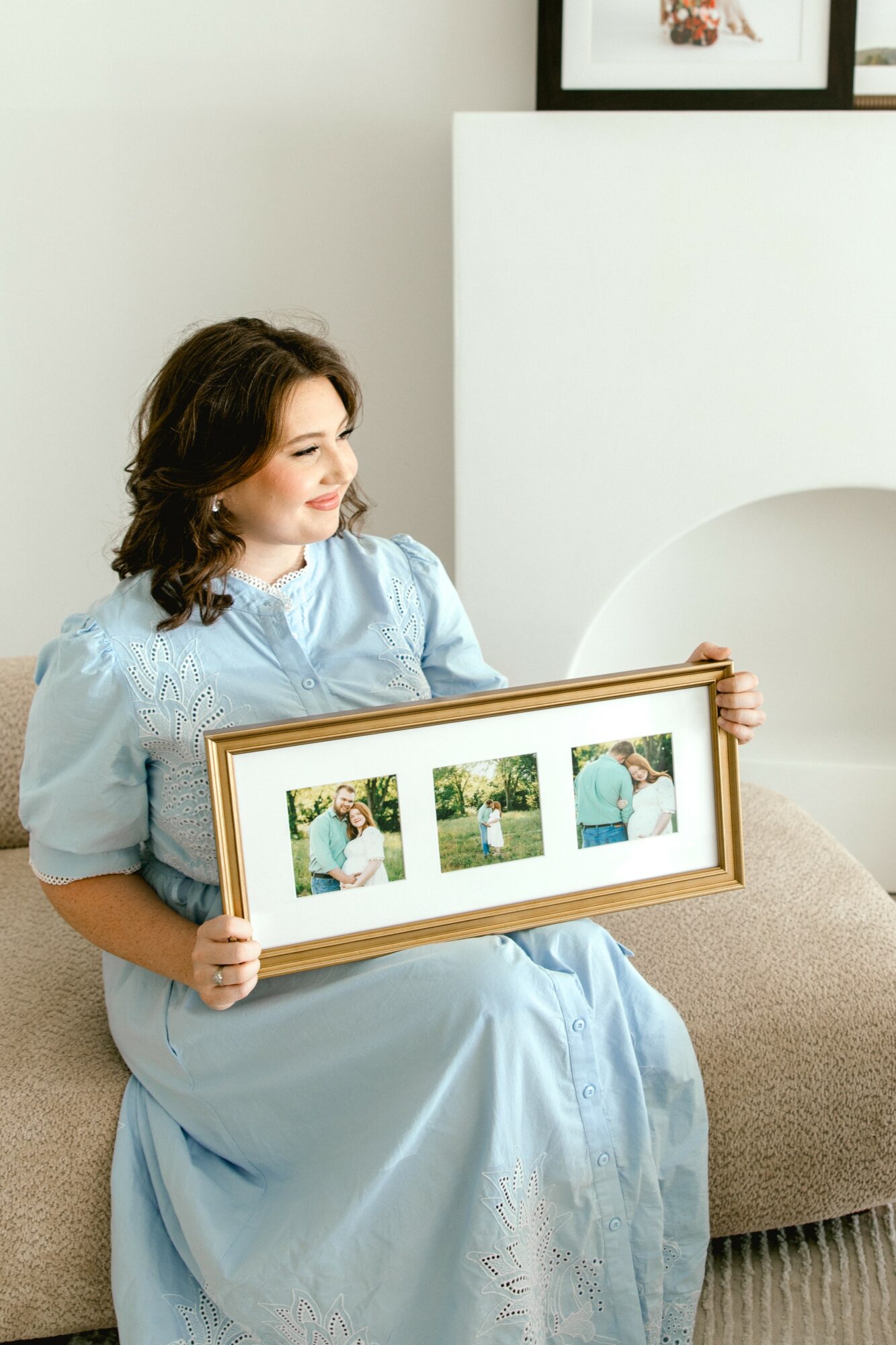 Woman sitting on a bench holding a framed collage of three photos, smiling, in a bright room.