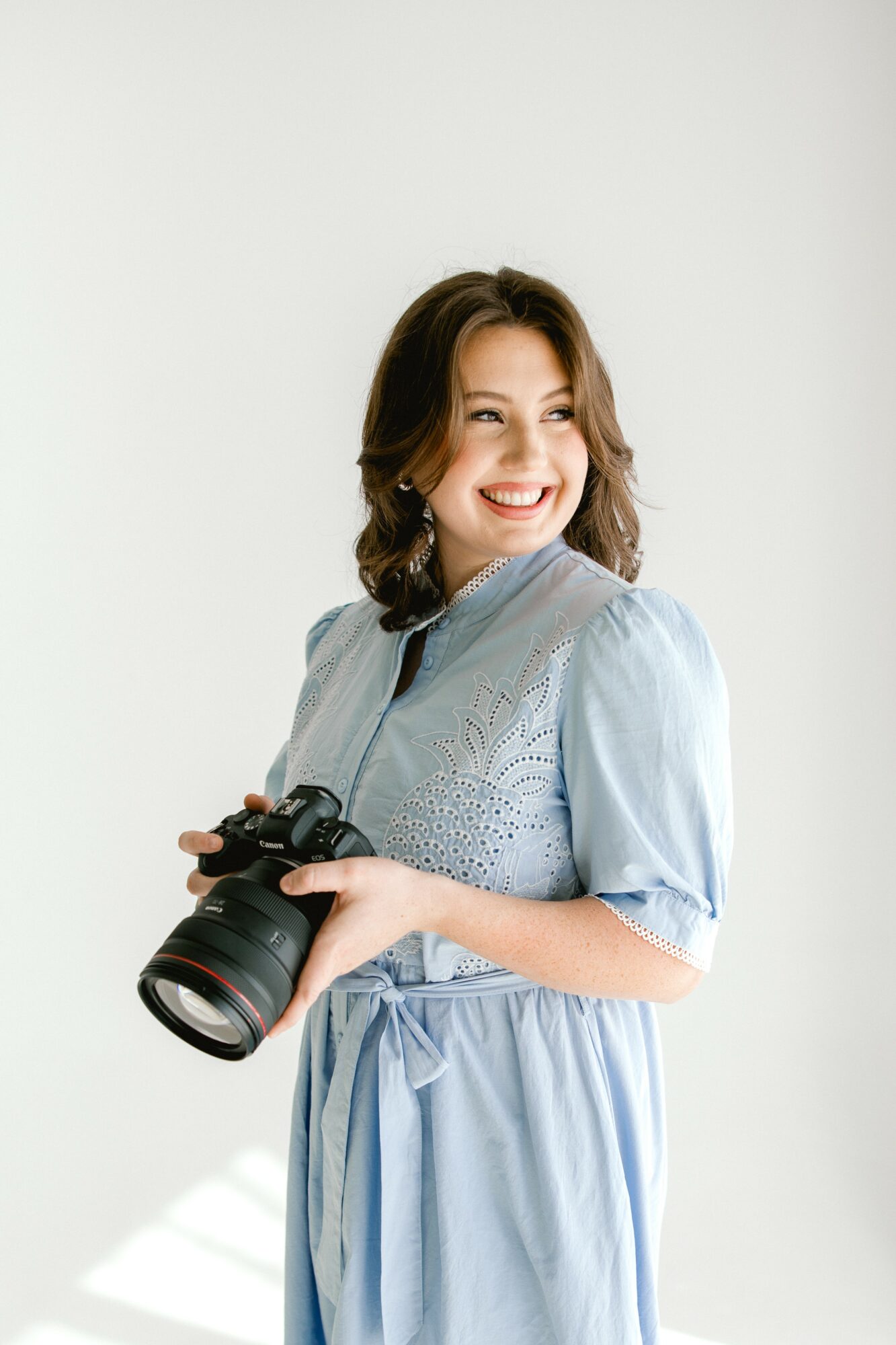 Young woman with shoulder-length hair smiling, holding a camera, wearing a light blue dress, standing against a plain background.