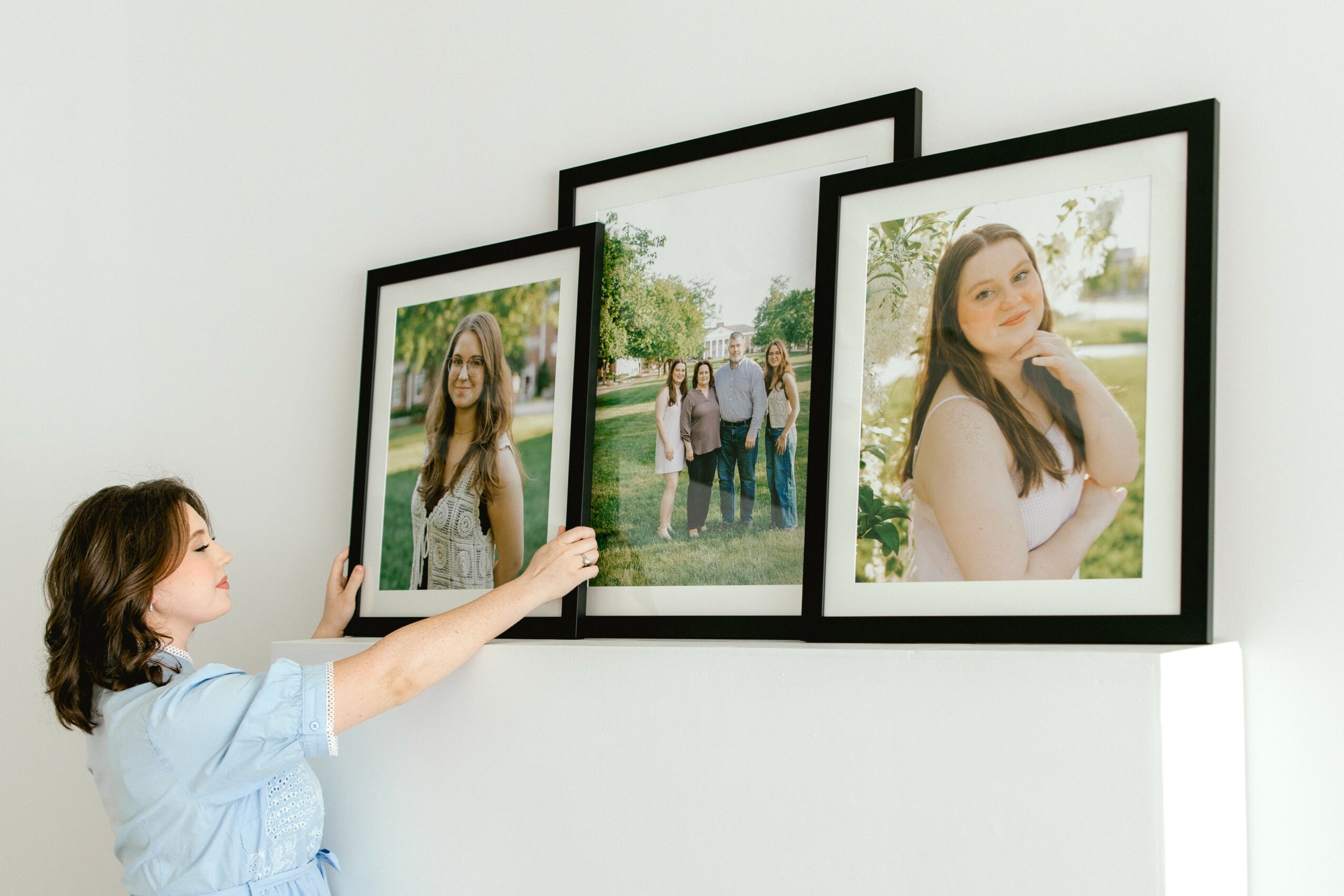 Child reaching towards framed photographs on a white wall, with a girl in a white dress in one photo, outdoors setting.