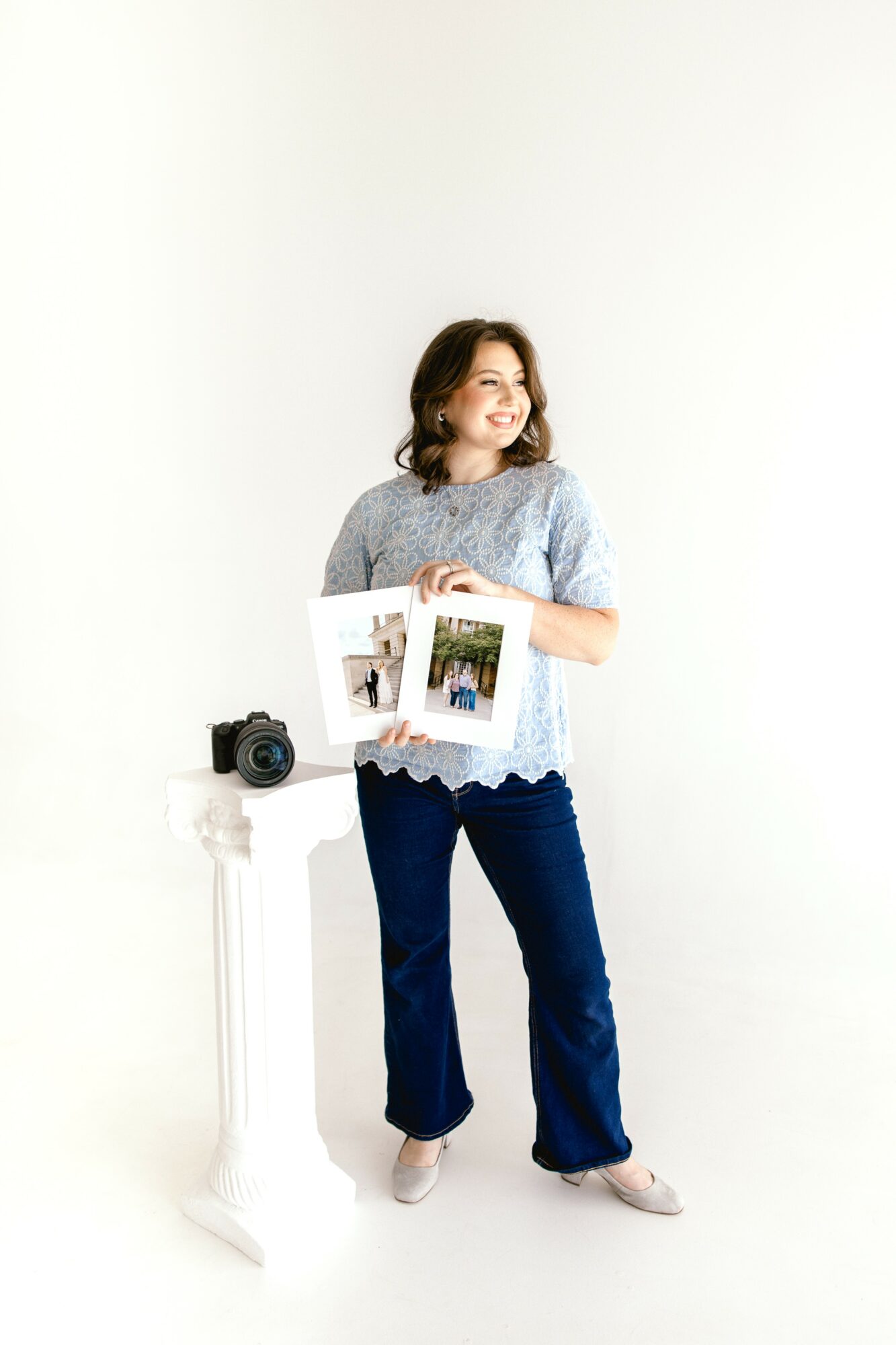 Woman holding photos, standing next to a small table with a camera, smiling, against a white background.