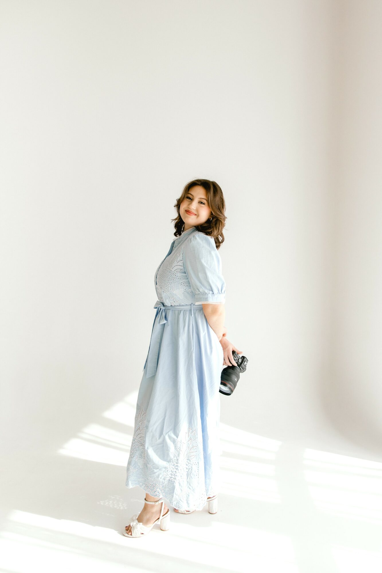 Woman with curly hair in a light blue dress holding a camera, standing in a bright, minimal space.