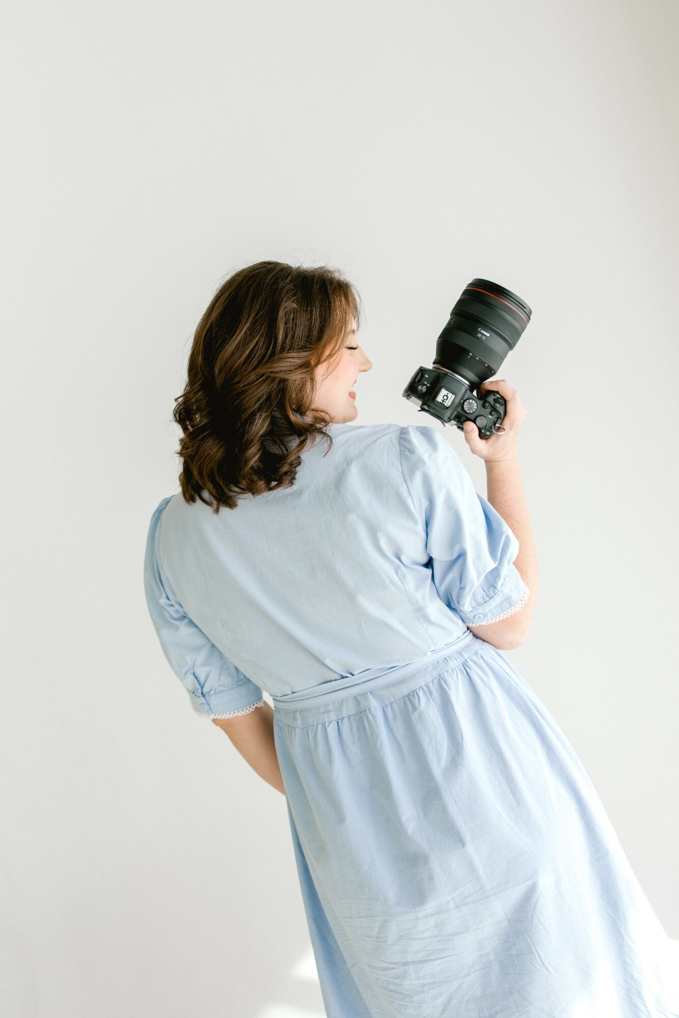 Woman with shoulder-length hair holding a camera with a large lens, facing sideways, against a plain white background.