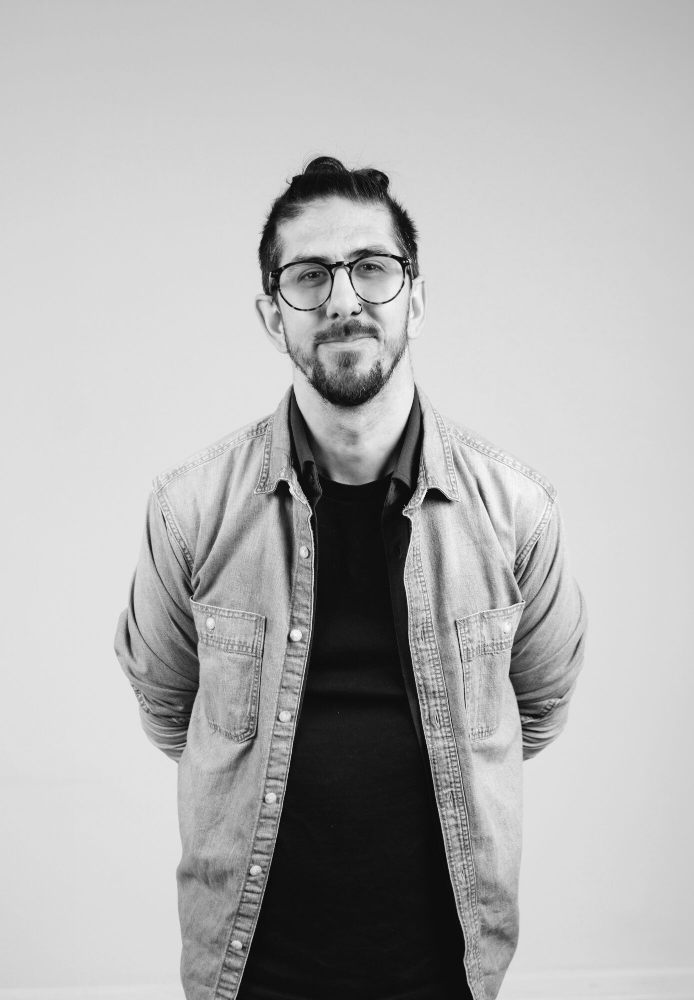 Man with glasses and a beard, wearing a denim jacket over a black shirt, standing against a plain background.