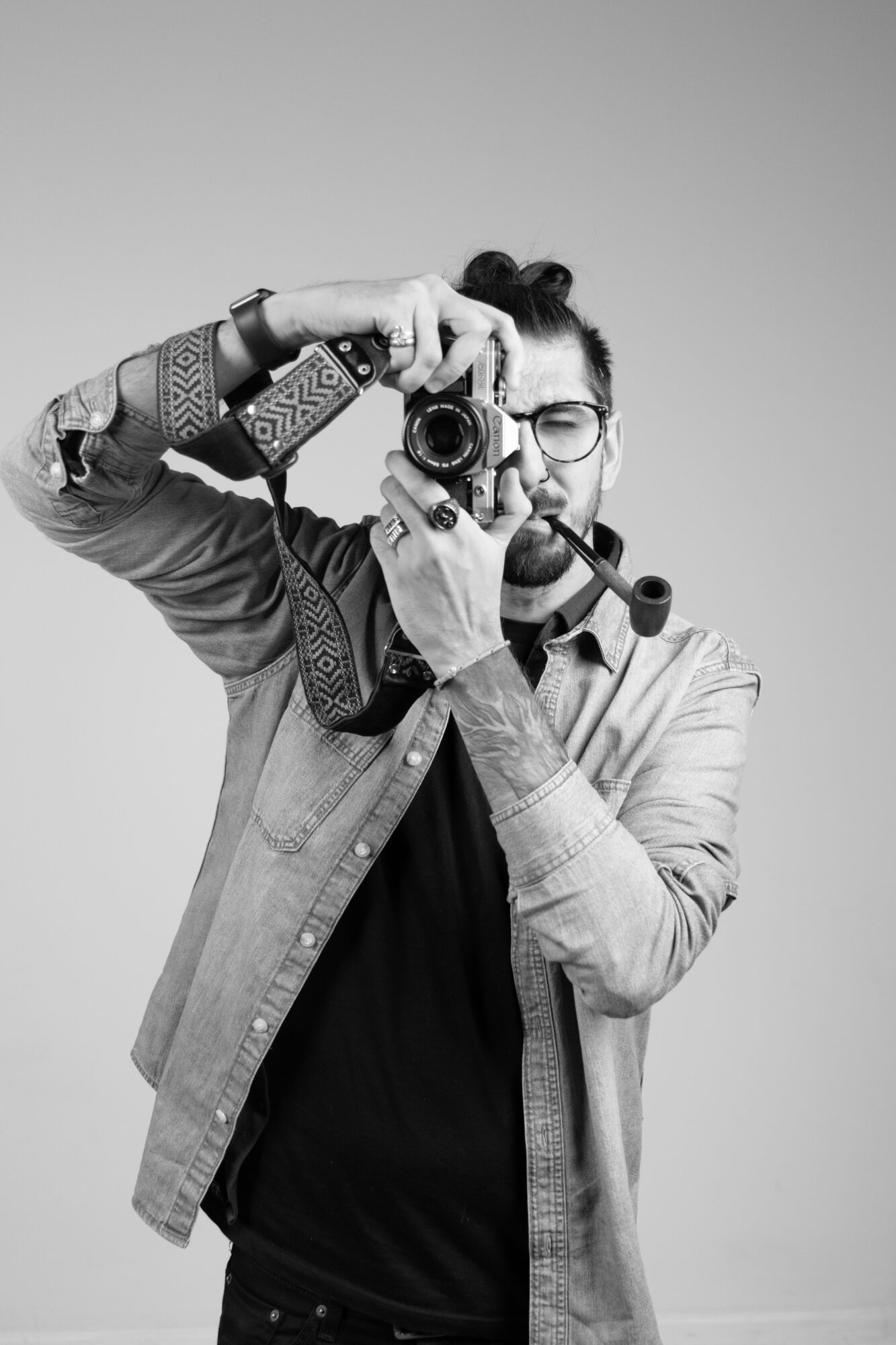 Person holding a camera, wearing glasses and a patterned jacket, taking a photo, with a plain background.