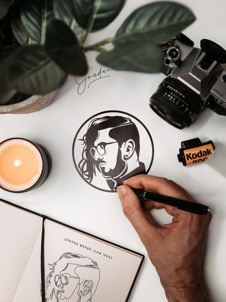 Person drawing a circular portrait of a man with glasses and a beard on paper, with a camera, candle, plant, and Kodak box nearby.