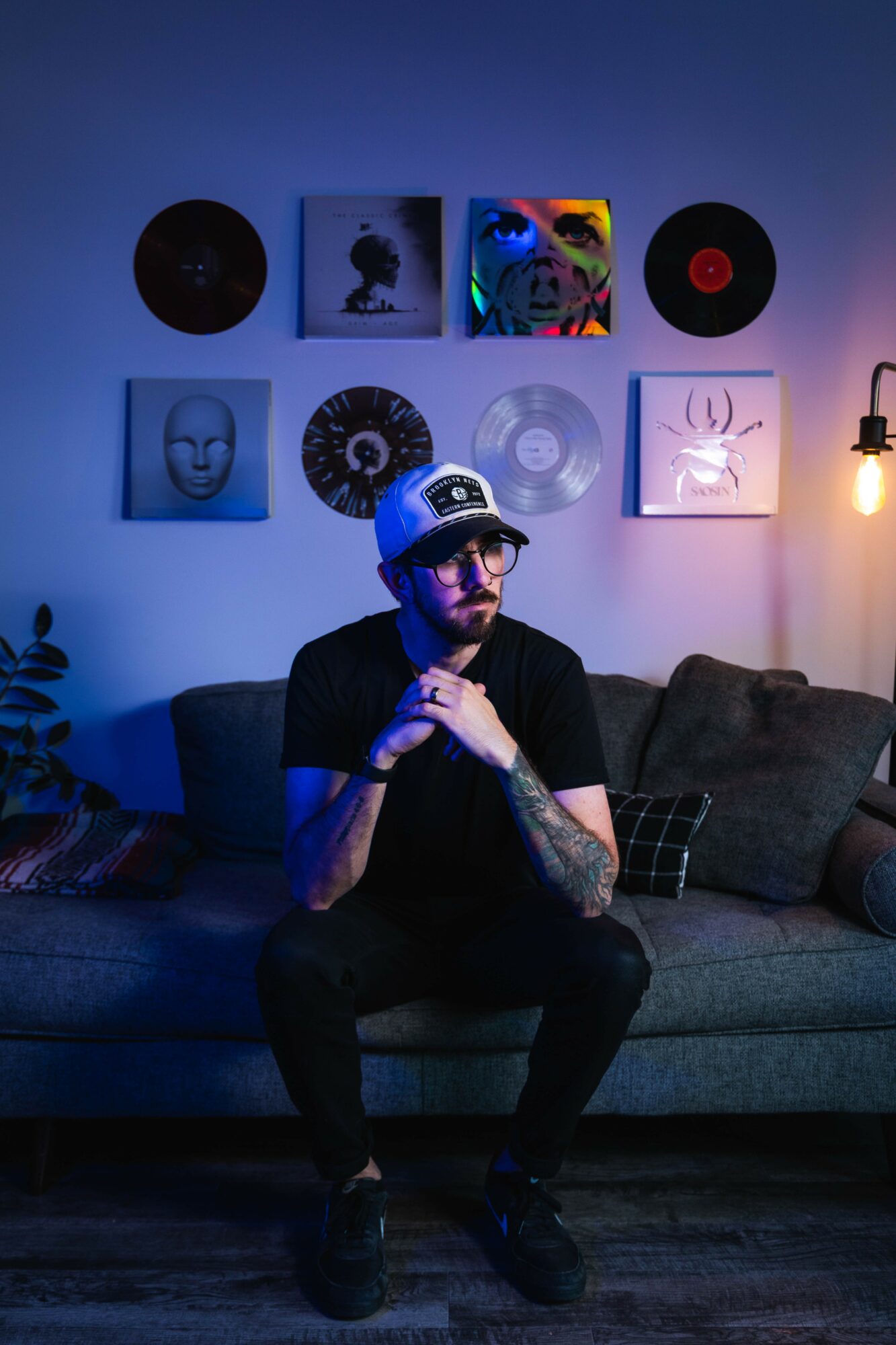 Man with glasses and a cap sitting on a sofa in a dimly lit room with wall art and a lamp.