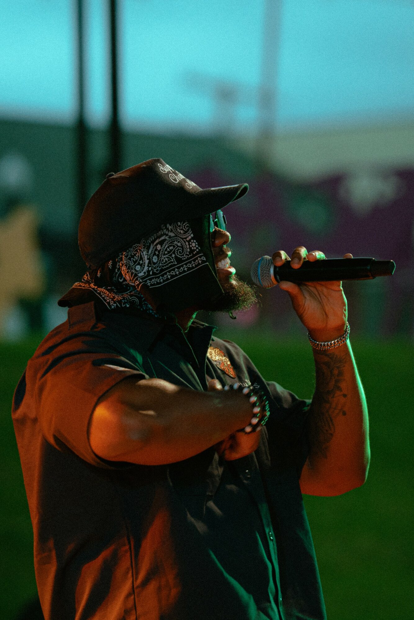 Person singing into microphone, wearing a bandana and cap, with a blurred outdoor background.