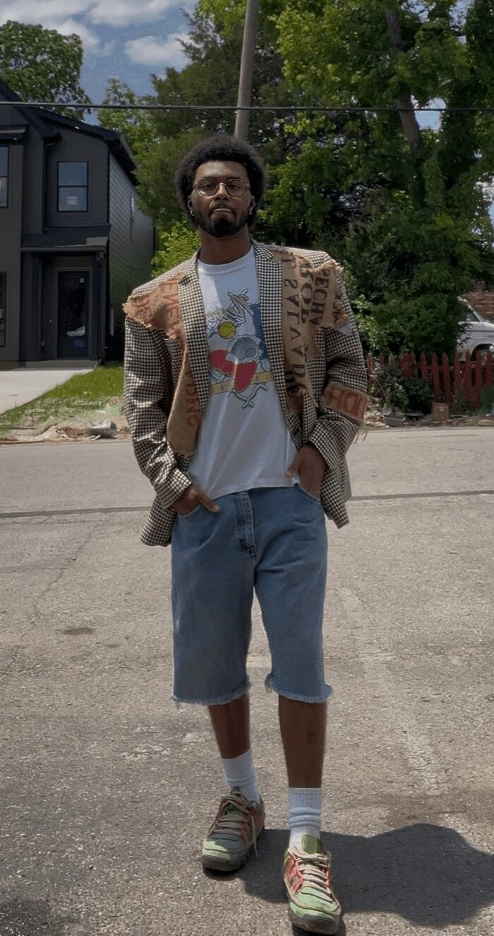 Young man standing outdoors on a street, wearing a beige jacket, white t-shirt, blue shorts, and sneakers.