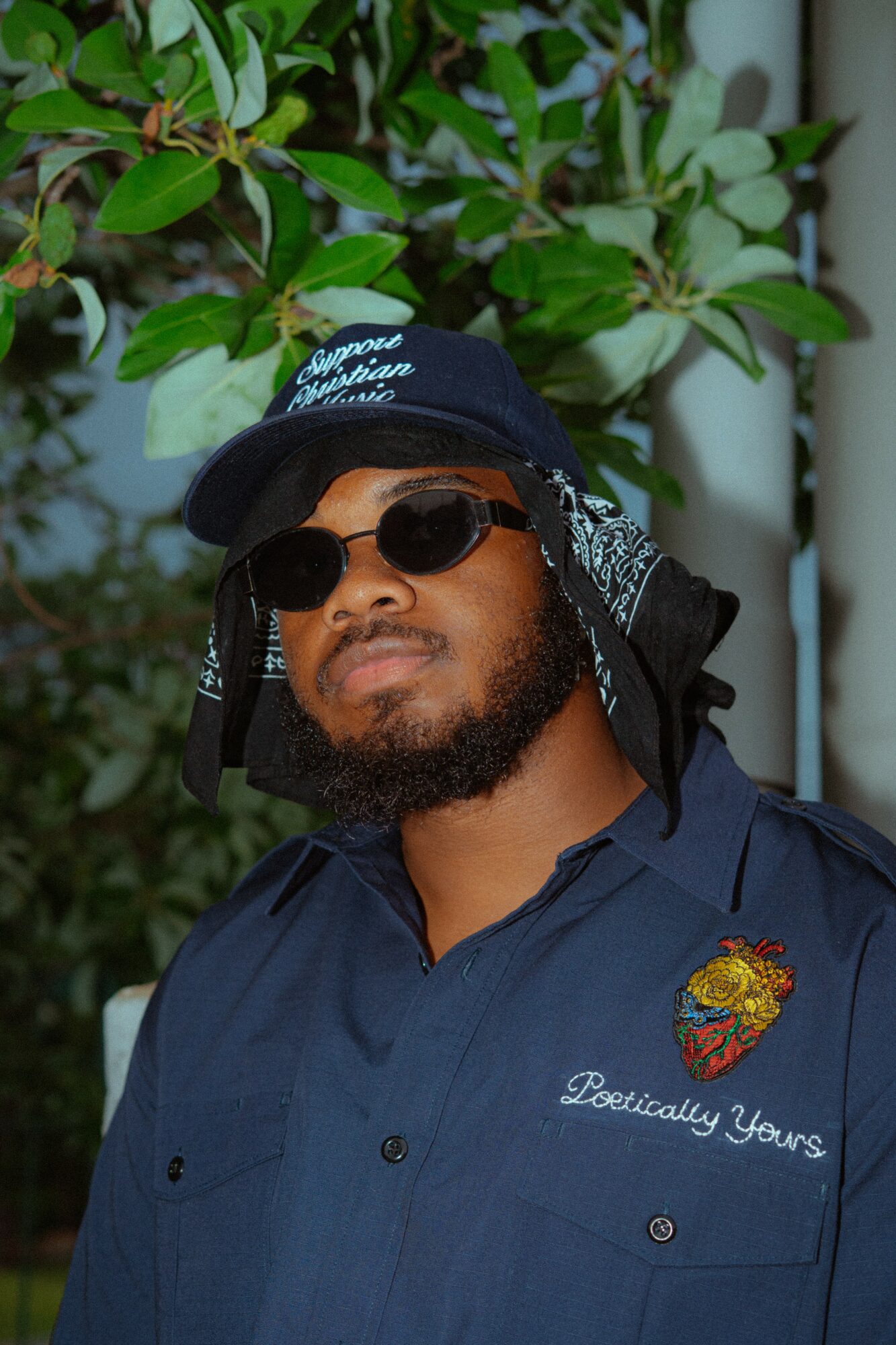 Man wearing sunglasses, a hat, and a navy shirt with embroidered patch, standing outdoors with green foliage behind him.