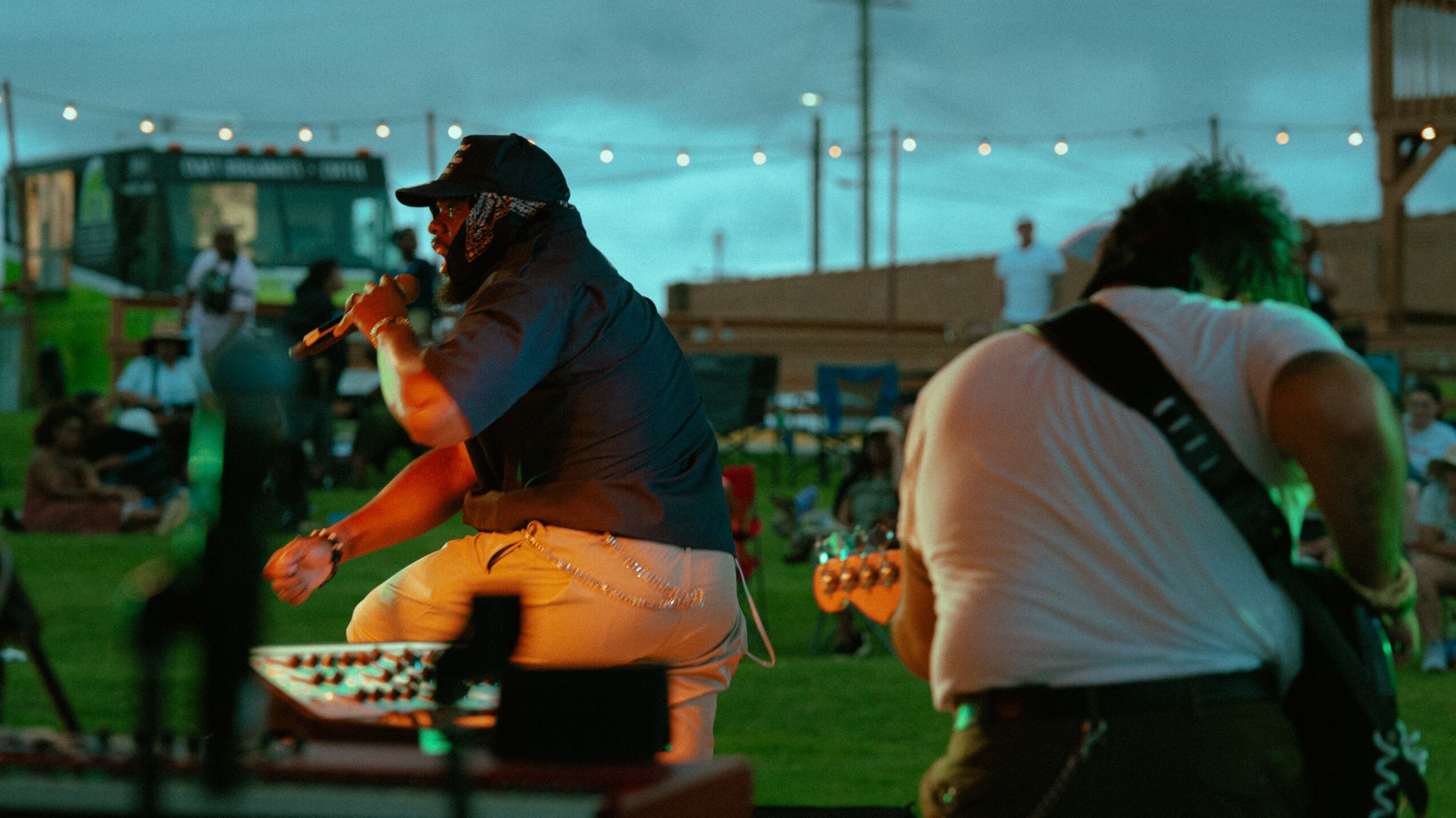 Two musicians perform outdoors at night with string lights, surrounded by people and tents, in a lively setting.