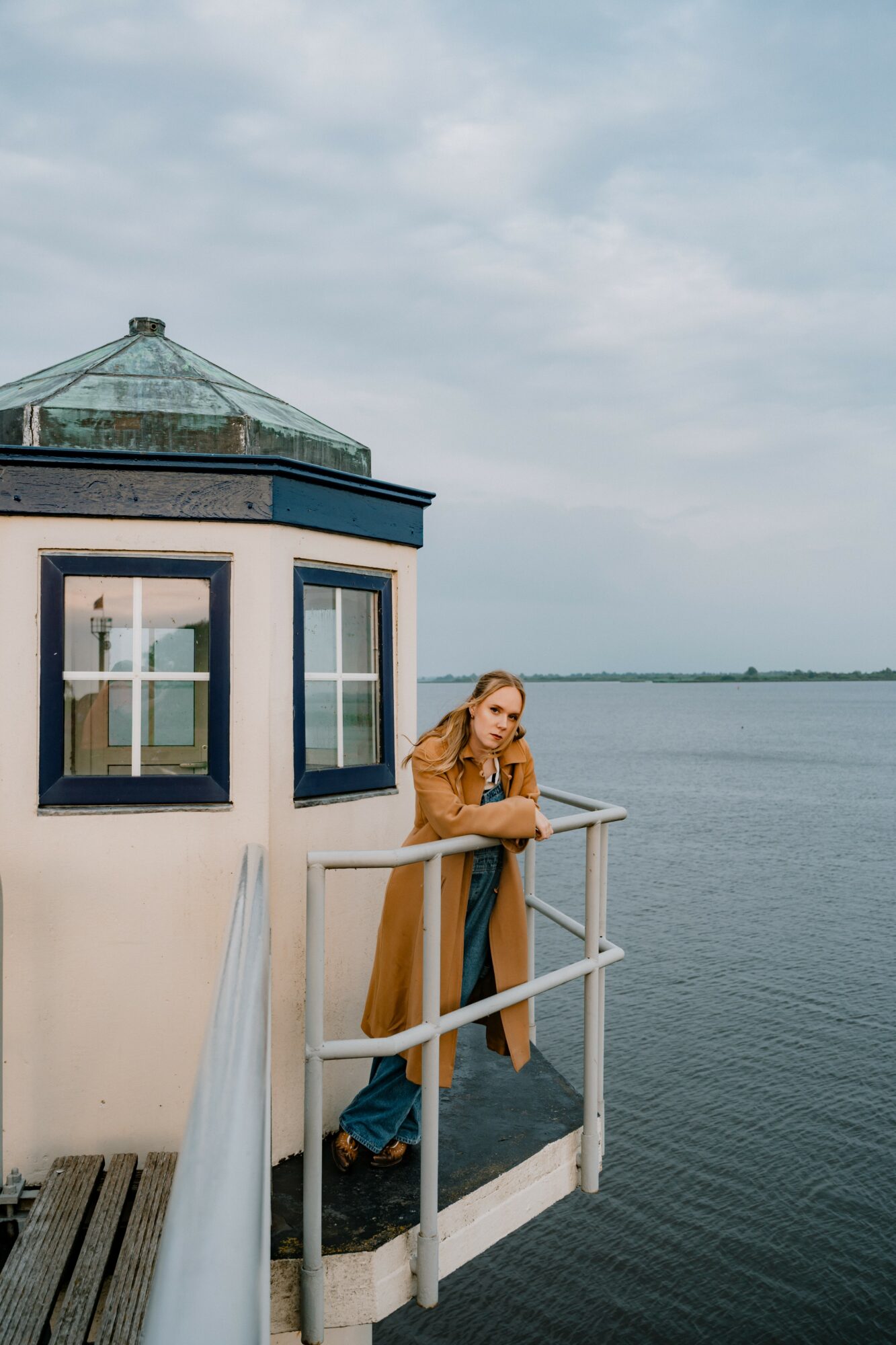 Woman in a brown coat stands on a dock near a small lighthouse by a body of water, looking at her phone.