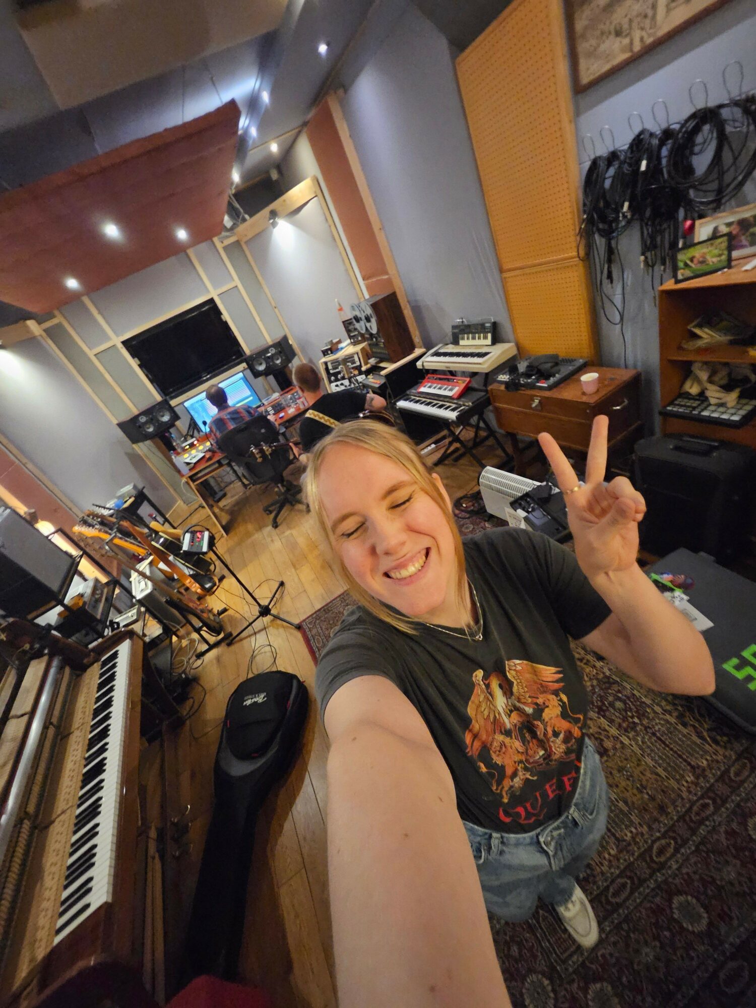 Person taking a selfie in a recording studio, smiling and making a peace sign, with musical equipment around.