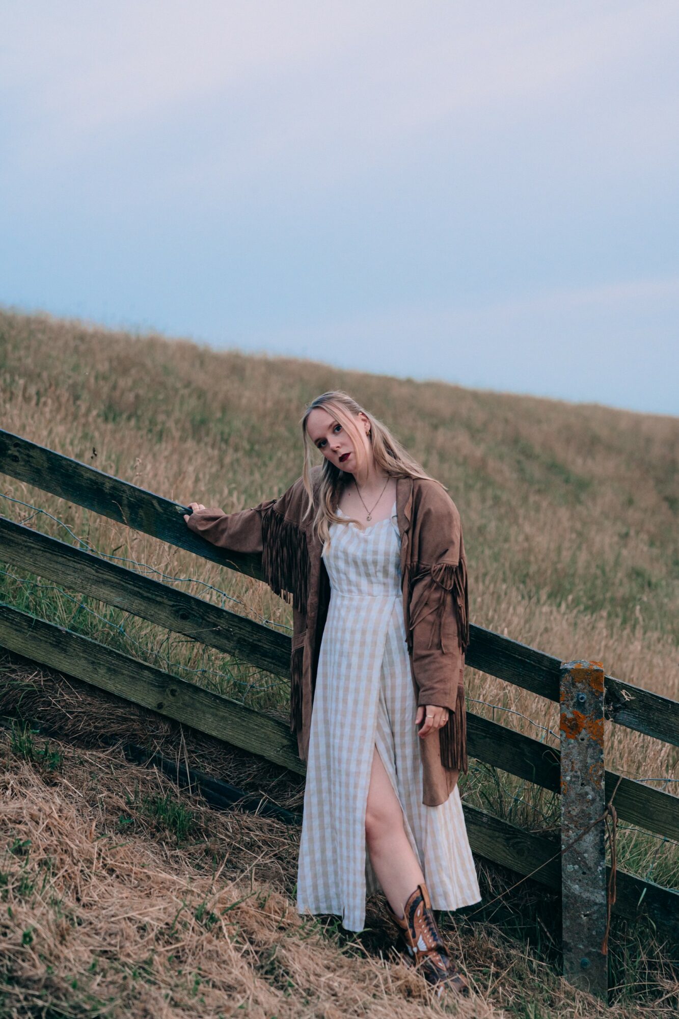 Woman in a white dress and brown coat standing by a wooden fence on a grassy hill, sky in background.