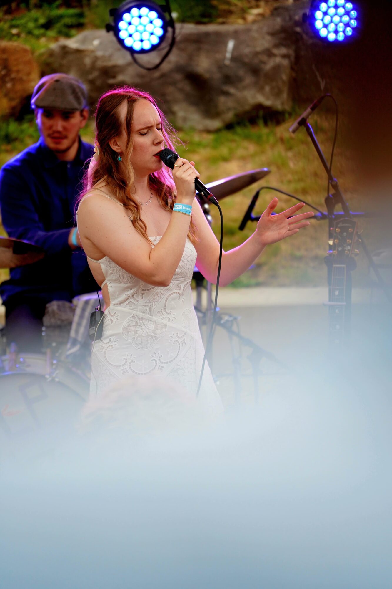 Young woman singing into a microphone with a person playing keyboard behind her outdoors, stage lights above.