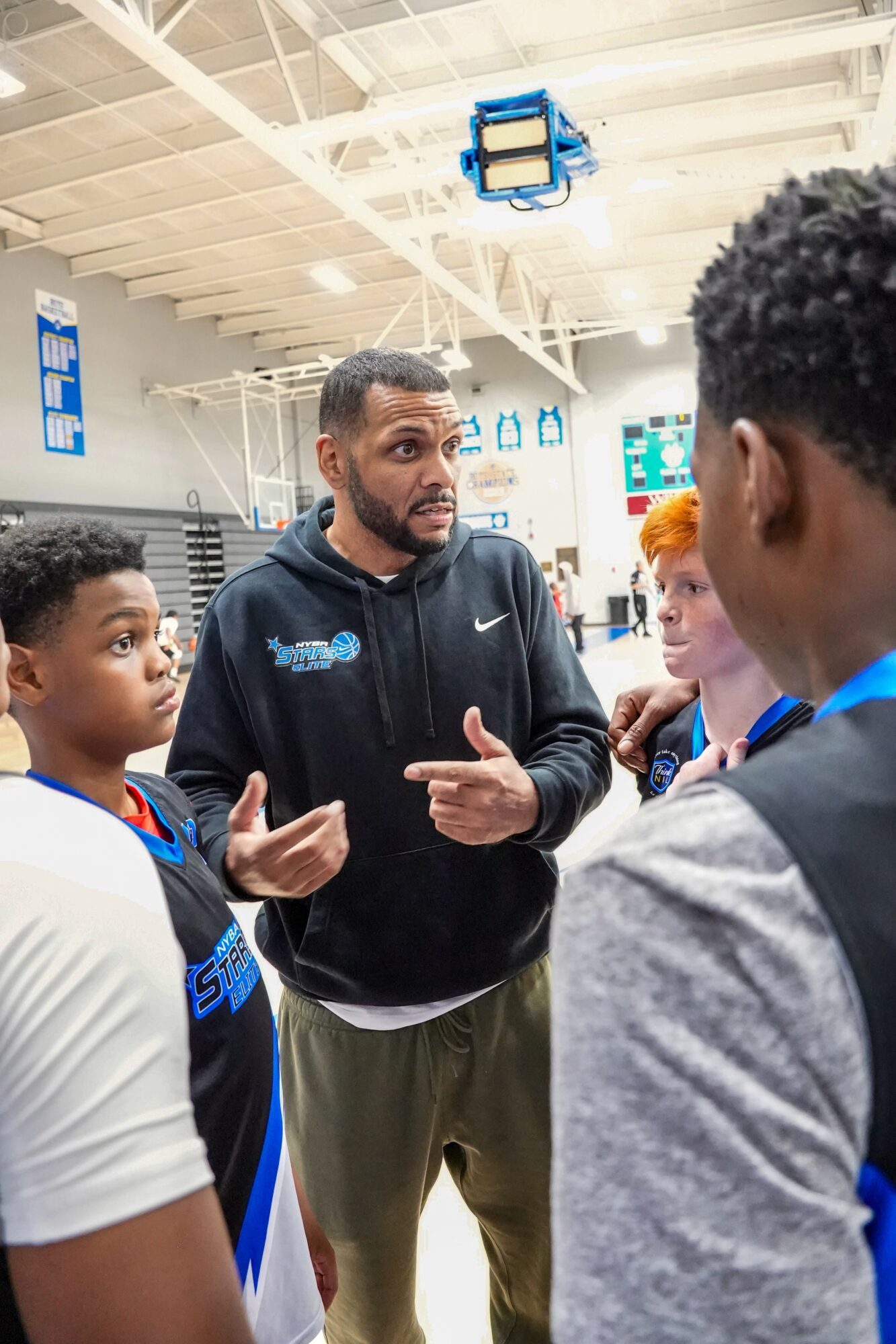Coach speaking to young basketball players in gymnasium, engaging in discussion, with ceiling lights and banners visible.
