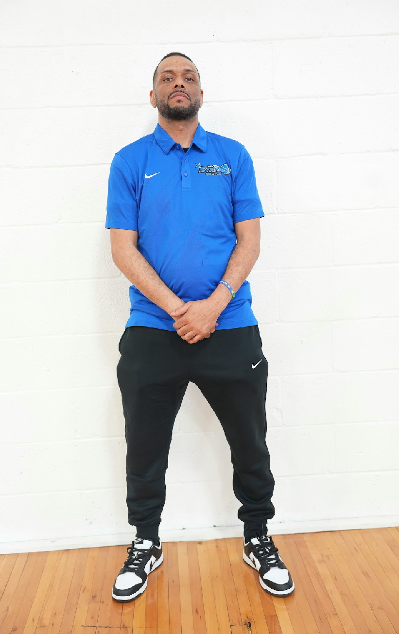 Young man standing with hands clasped, wearing a blue polo shirt, black pants, and sneakers, against a white wall.