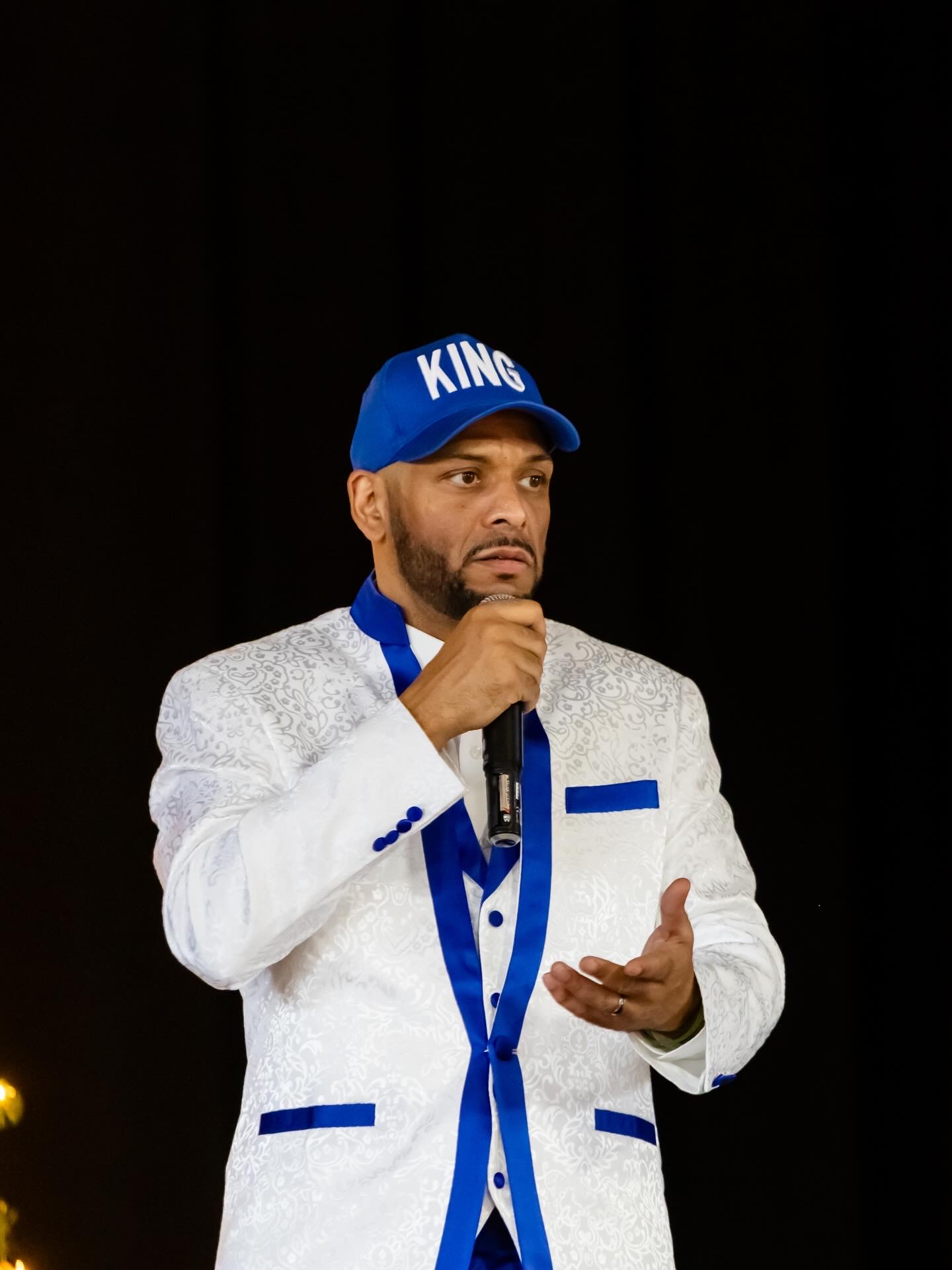 Man wearing a blue cap with 'KING' and a white suit with blue accents, speaking into a microphone, against a black background.