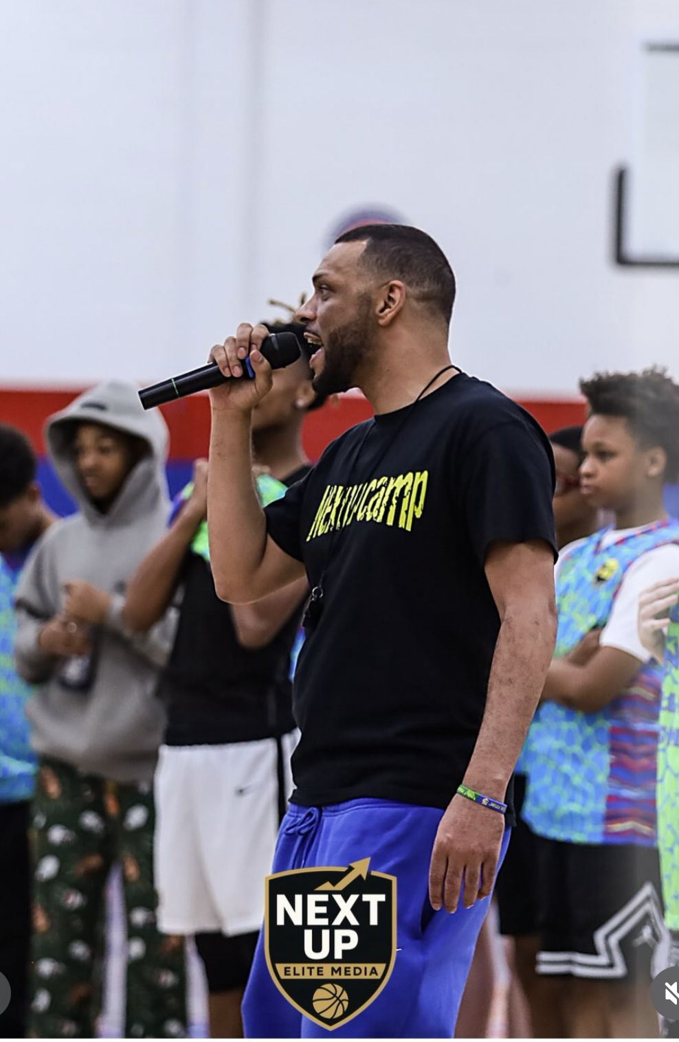 Man speaking into microphone at indoor event with children in background, some wearing colorful shirts.