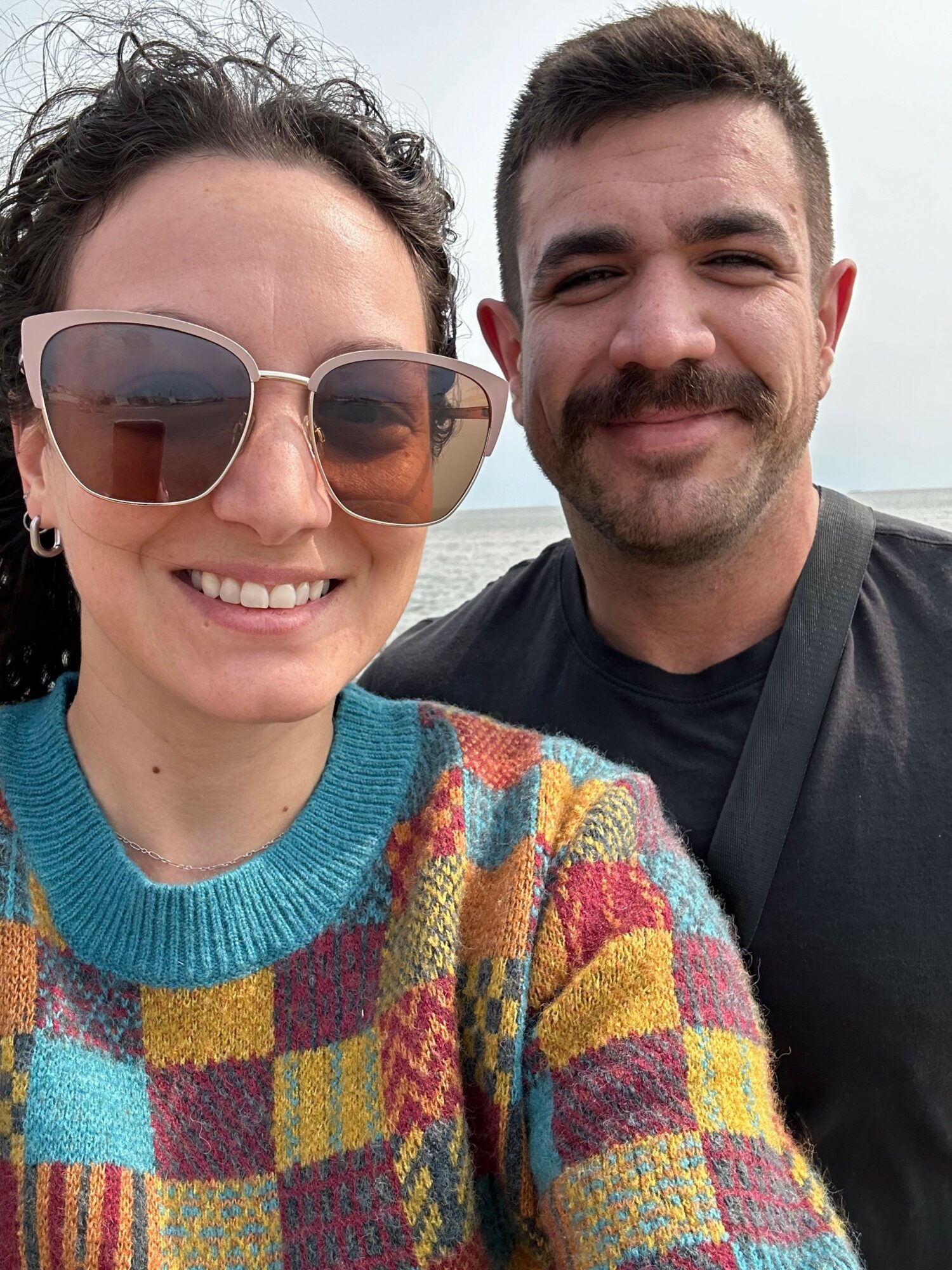 Two smiling people, one wearing sunglasses, close together outdoors with a colorful blanket or towel, smiling at the camera.