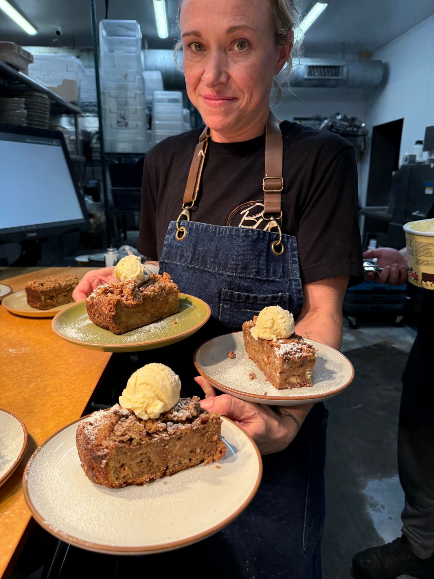 Person holding a plate with cake and ice cream, standing in a kitchen. Two plates with cake and ice cream are on the table.