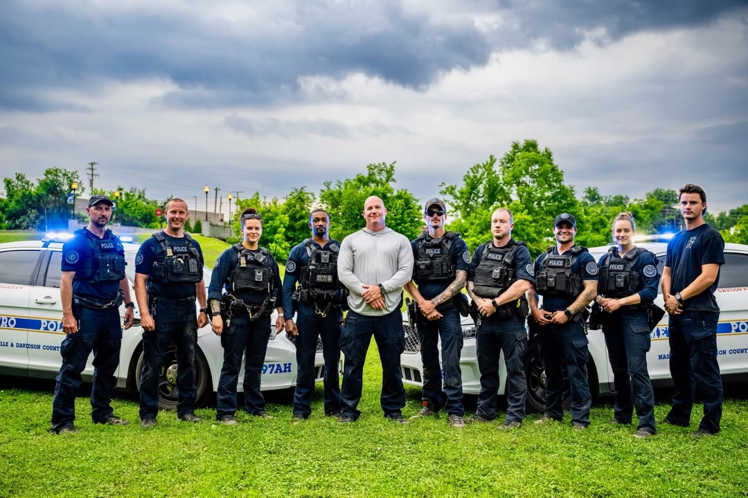 Group of nine police officers and one civilian standing outdoors in front of police cars, with trees and cloudy sky behind them.