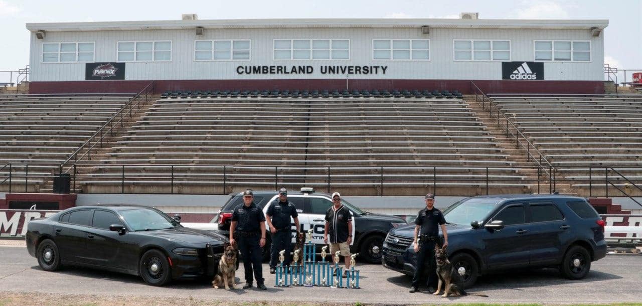 Group of people standing between two black cars in front of a stadium with empty bleachers and a Cumberland University sign.