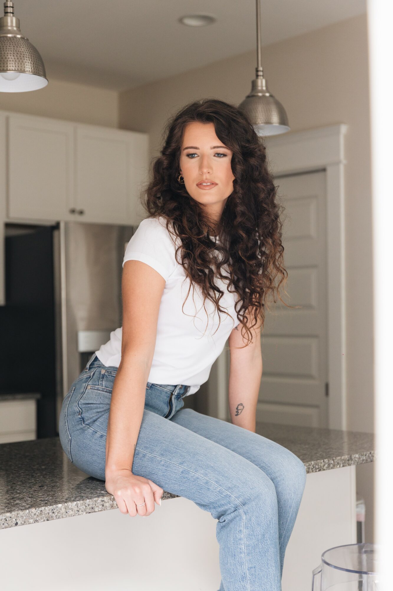 Young woman with curly hair sitting on kitchen counter, wearing a white t-shirt and jeans, in a modern kitchen.