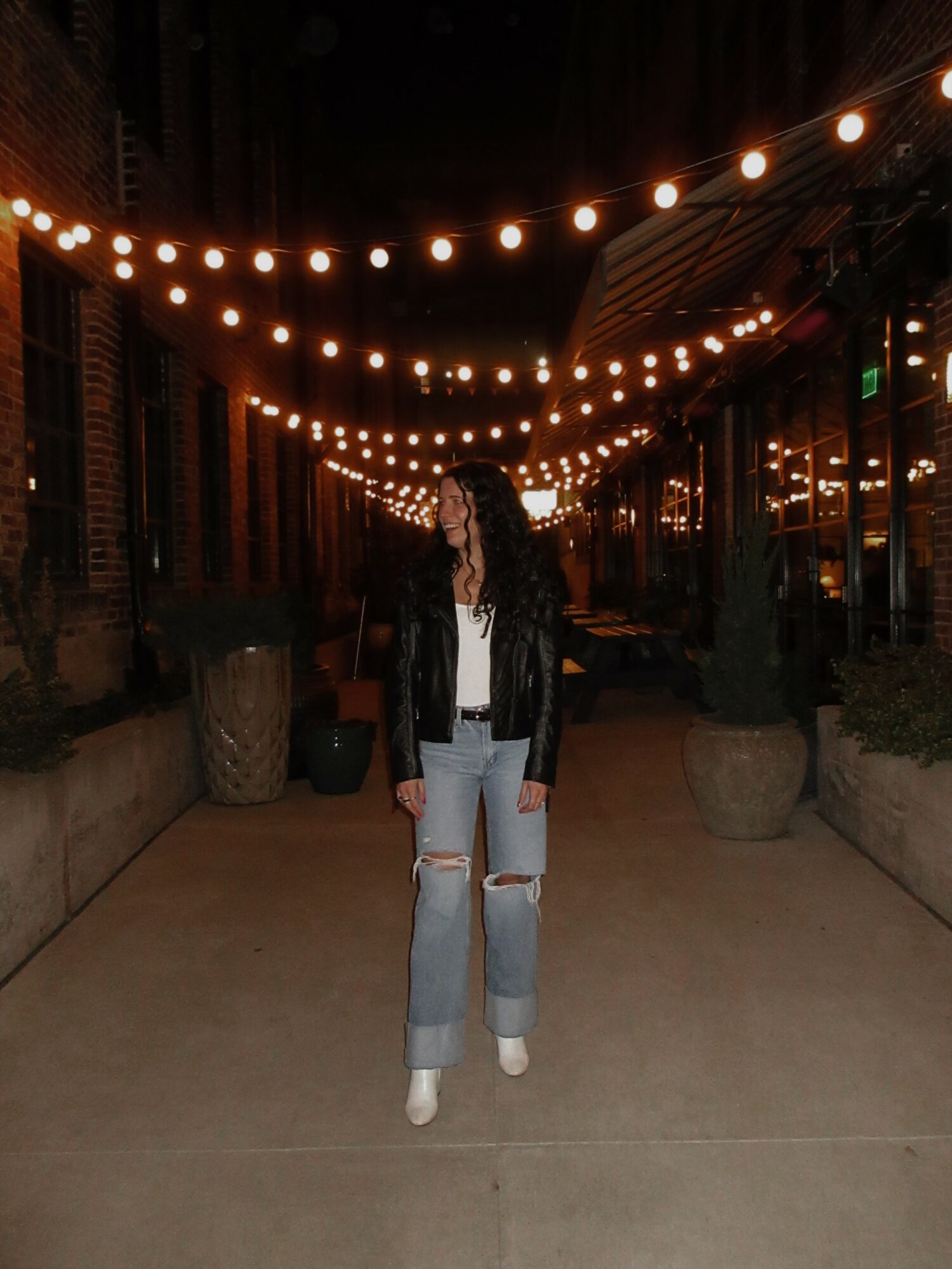 Person standing on a lit outdoor street at night with string lights overhead, wearing a black jacket and ripped jeans.