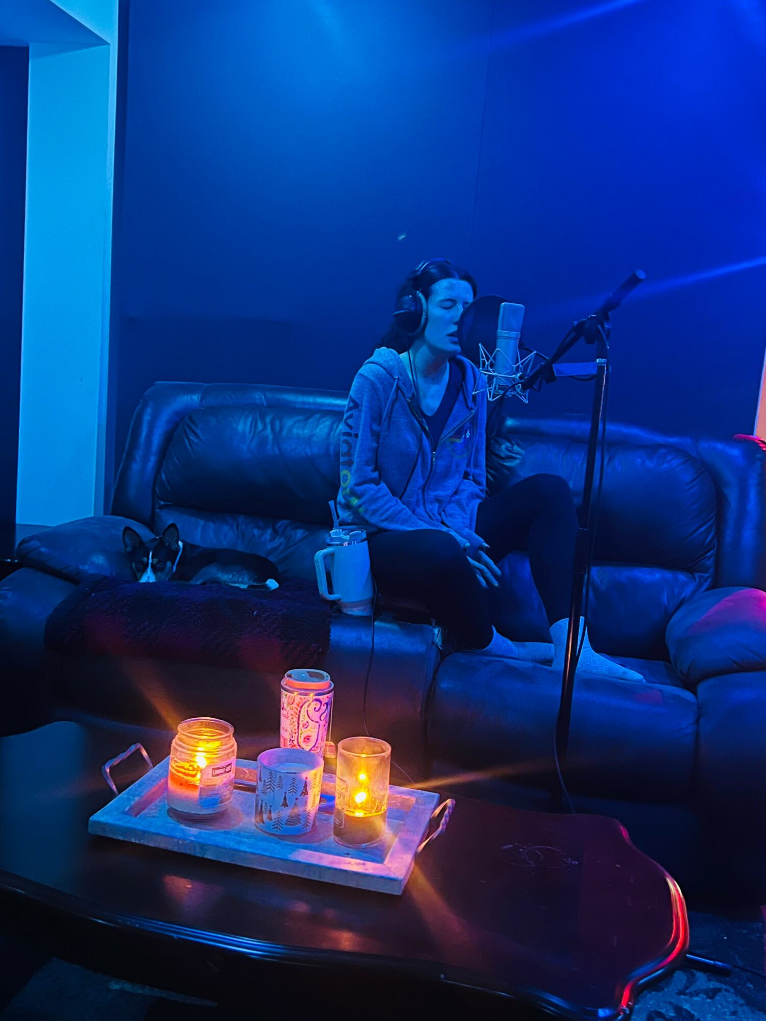 Woman sitting on a black leather couch in a dimly lit room with blue lighting, recording vocals with a microphone, candles on a tray in front.