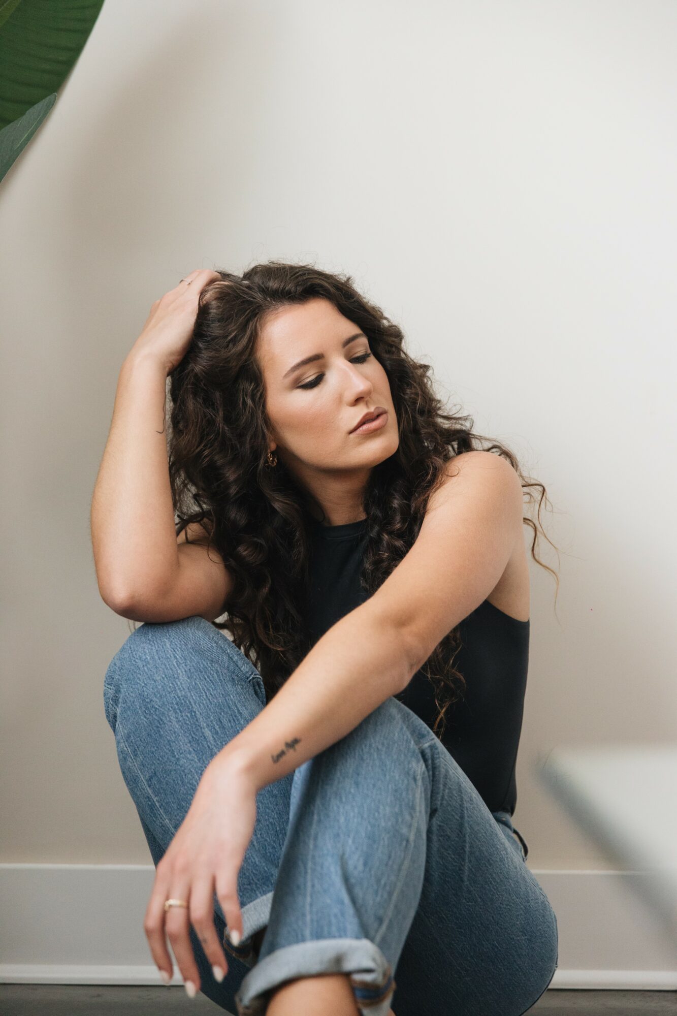 Young woman with curly hair sitting on the floor, resting her head on her hand, eyes closed, wearing a black top and jeans.