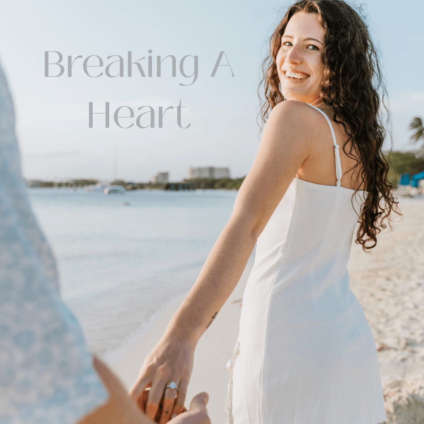 Smiling woman in white dress holding hands on beach with water and buildings in background
