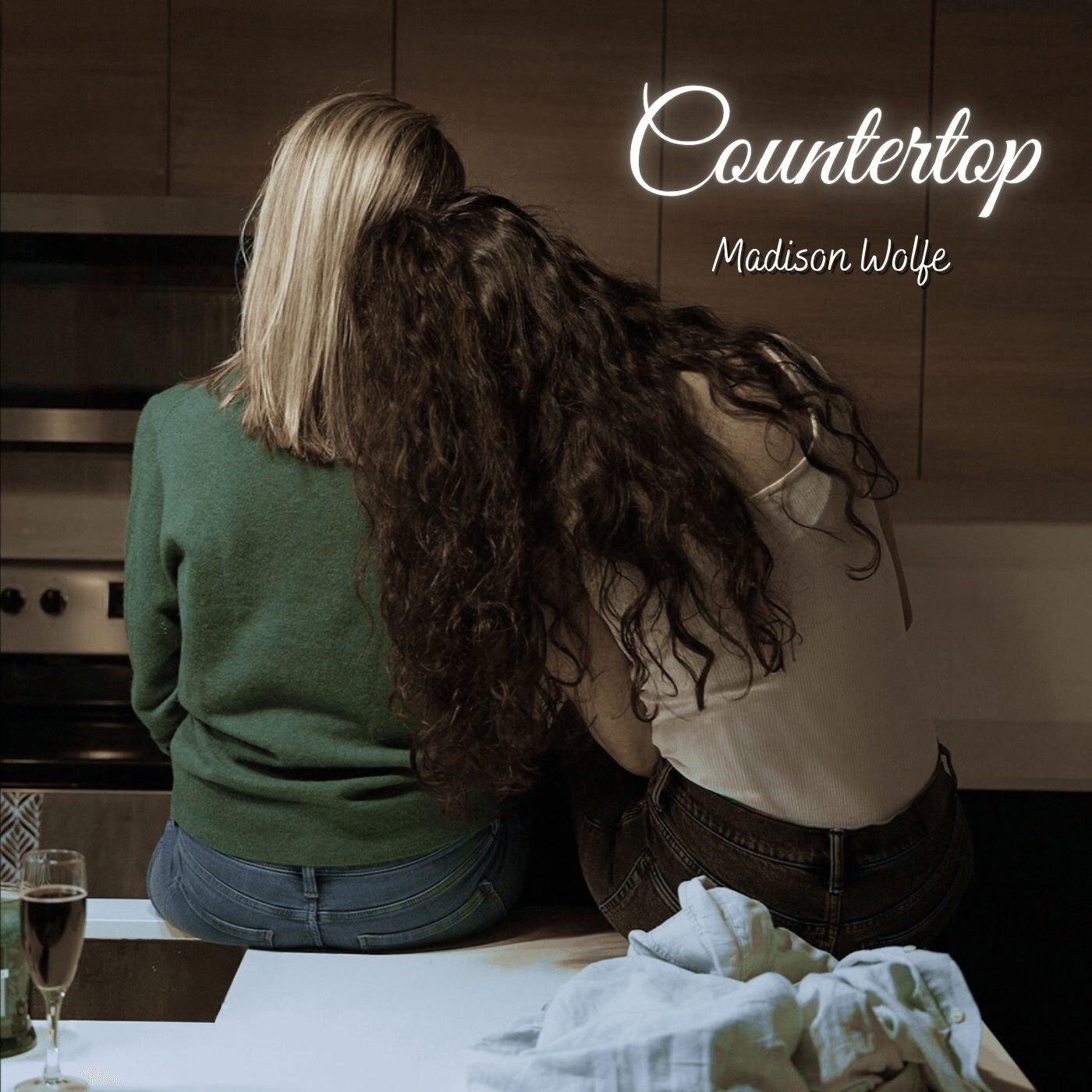 Two women sit on a kitchen counter, one with blonde hair and the other with long, curly dark hair, facing away.