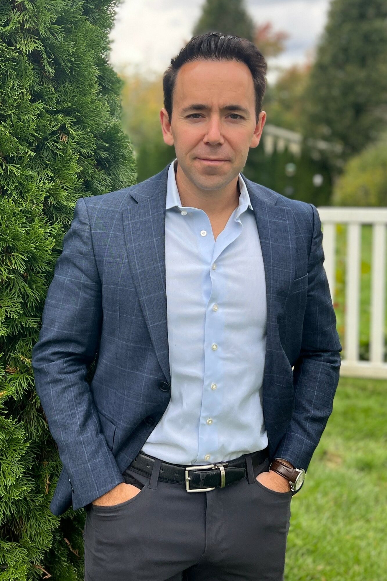Man in a blue checkered blazer and light blue shirt standing outdoors with greenery and a white fence behind him.