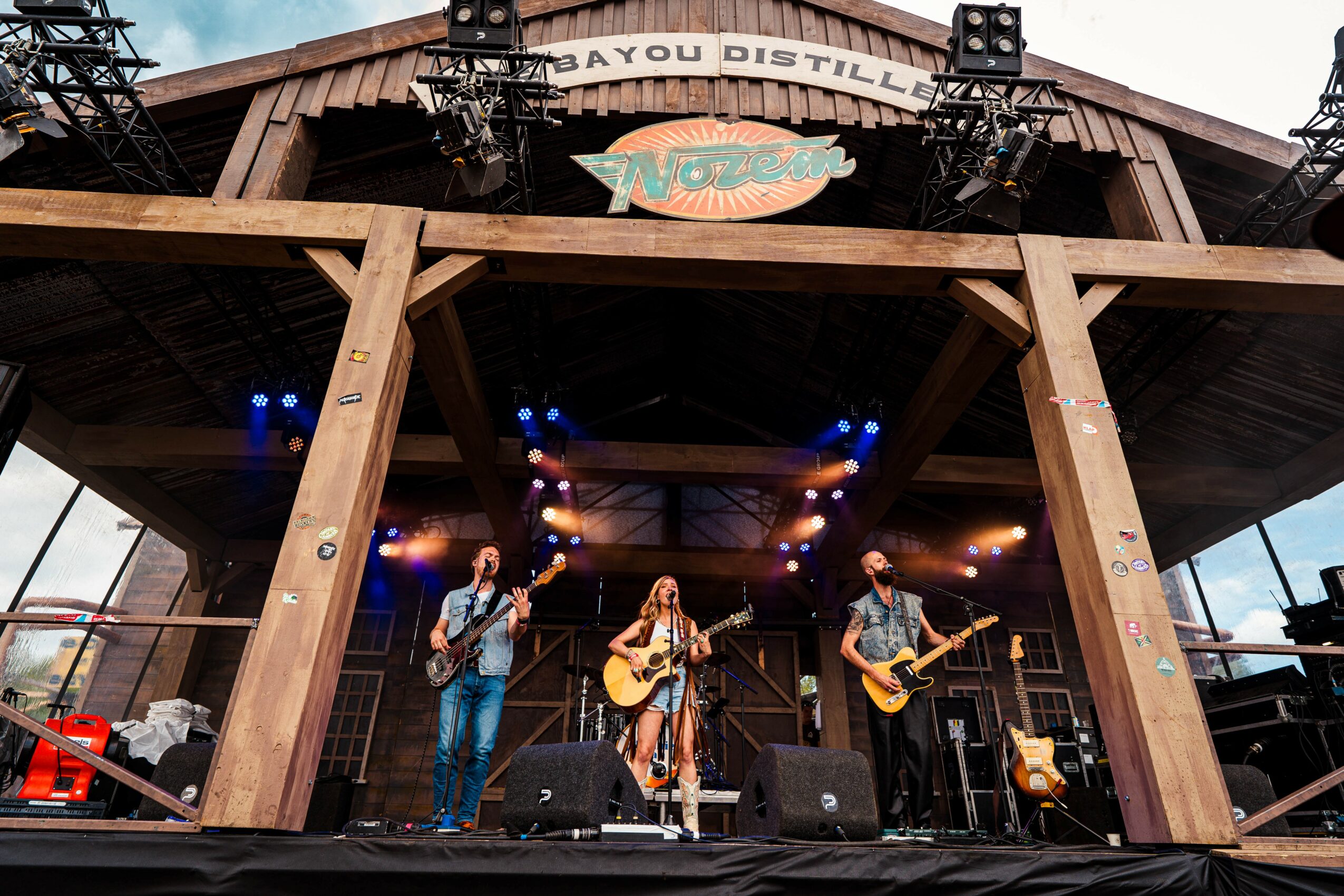 Three musicians perform on an outdoor stage with wooden beams, colorful lights, and a sign reading 'Bayou Distillery'.