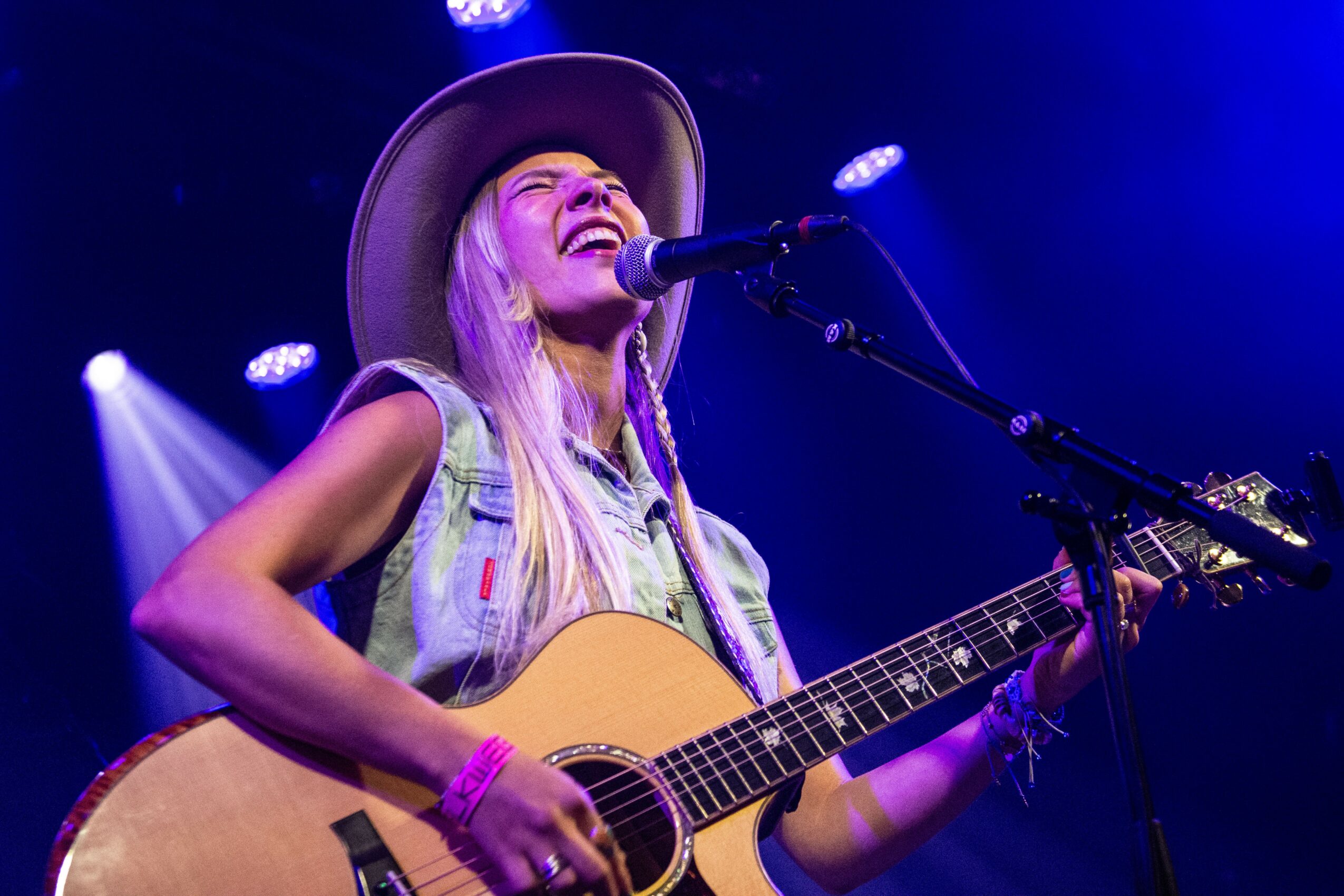 A woman with long blonde hair wearing a wide-brimmed hat plays an acoustic guitar and sings into a microphone on stage.