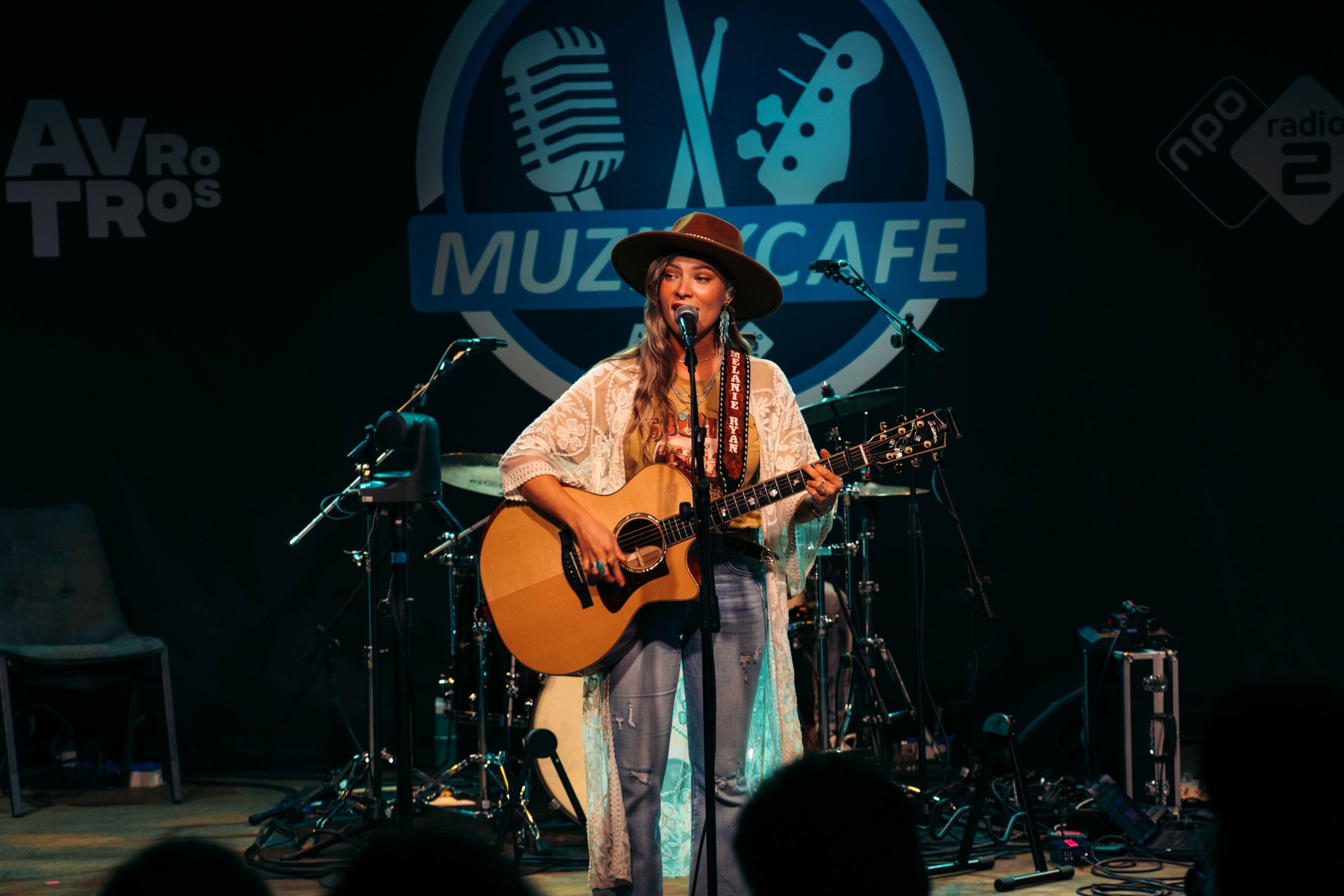 Female musician playing guitar and singing on stage with music equipment and a logo backdrop.