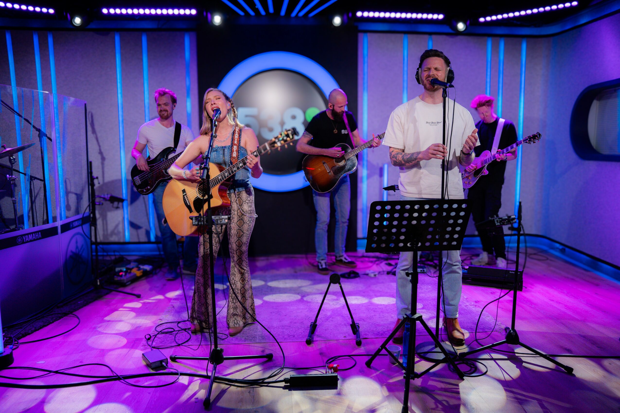 Band performing on stage with purple and blue lighting, two singers with guitars, and a screen in the background.