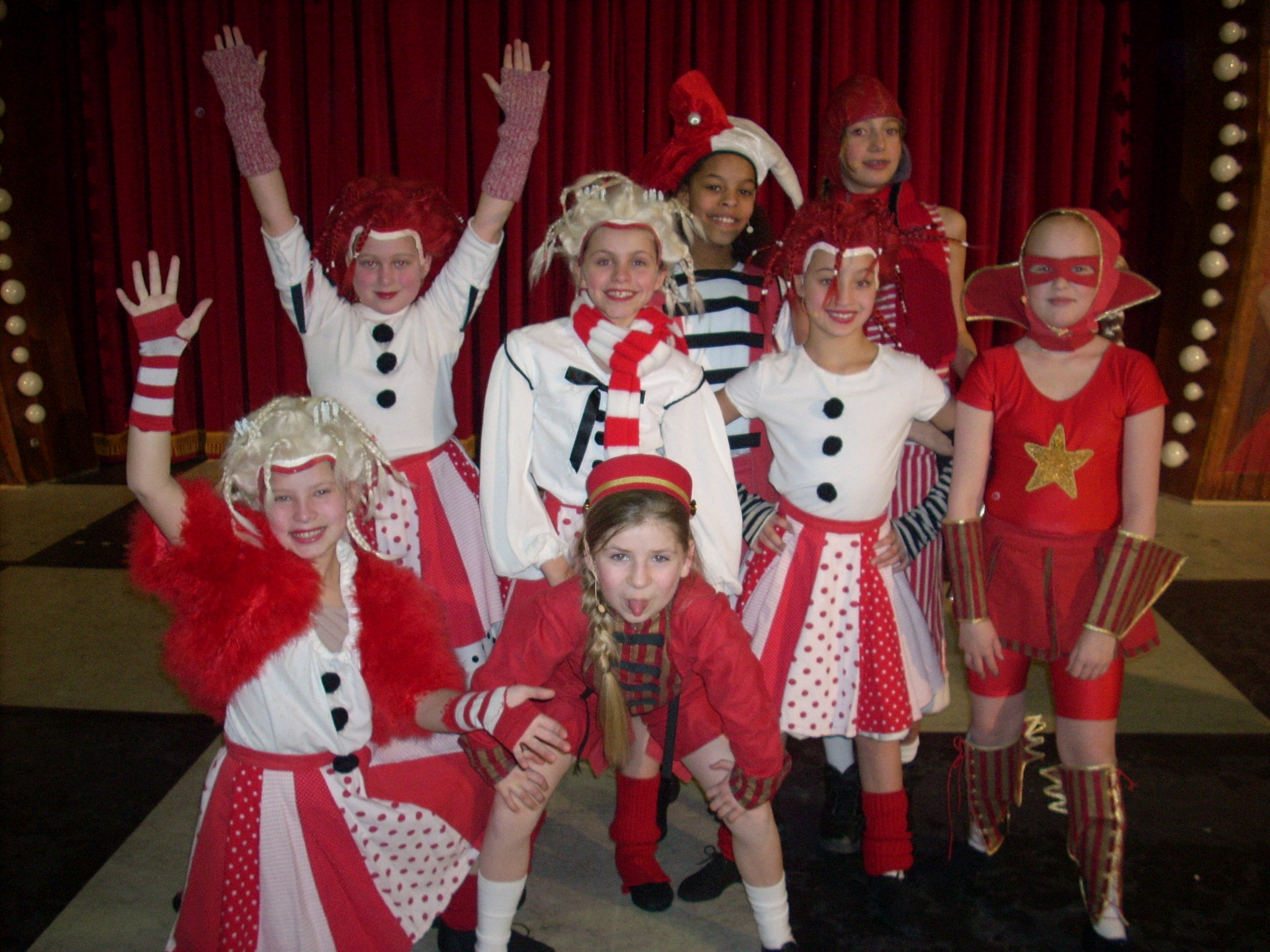 Group of children dressed in Christmas-themed costumes, standing on stage with red curtains in background.