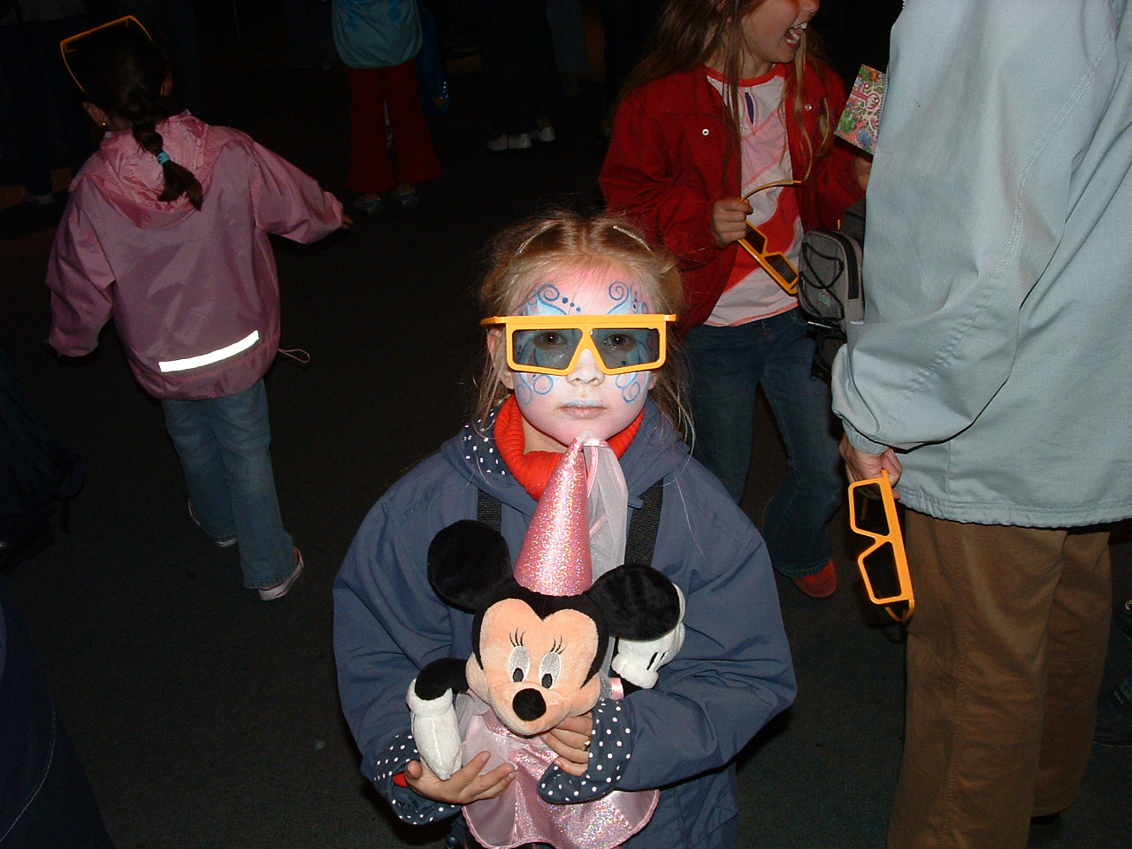 Child wearing sunglasses and a pink cone hat, holding Mickey Mouse plush toy, surrounded by other children and adults.