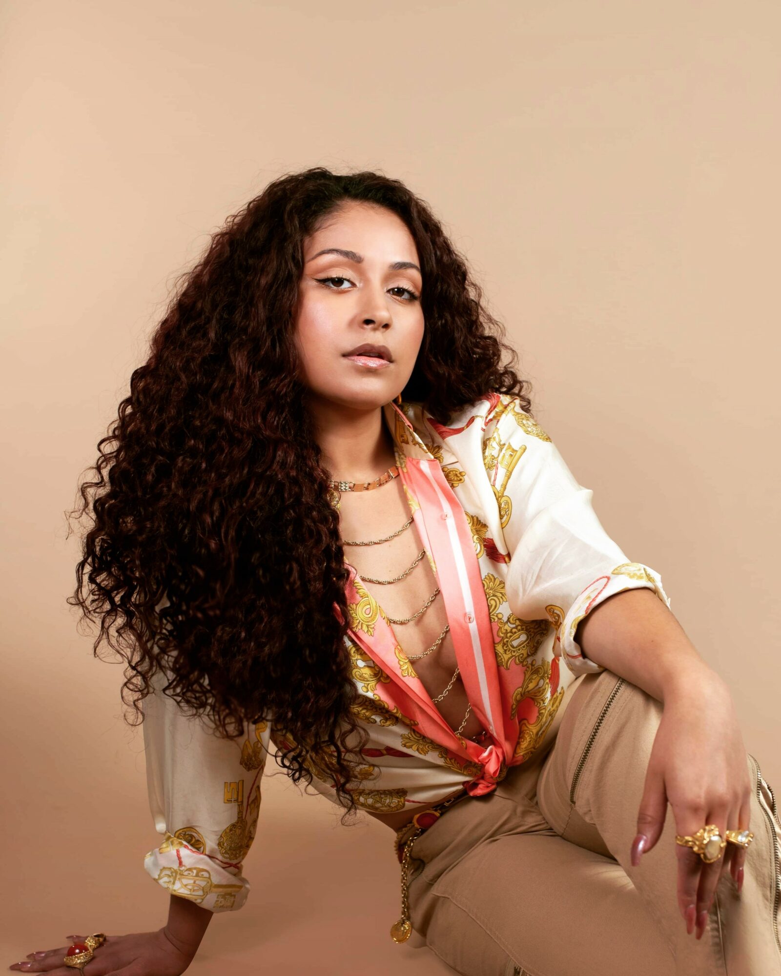 Young woman with long curly hair, wearing a patterned blouse and beige pants, sitting against a beige background.