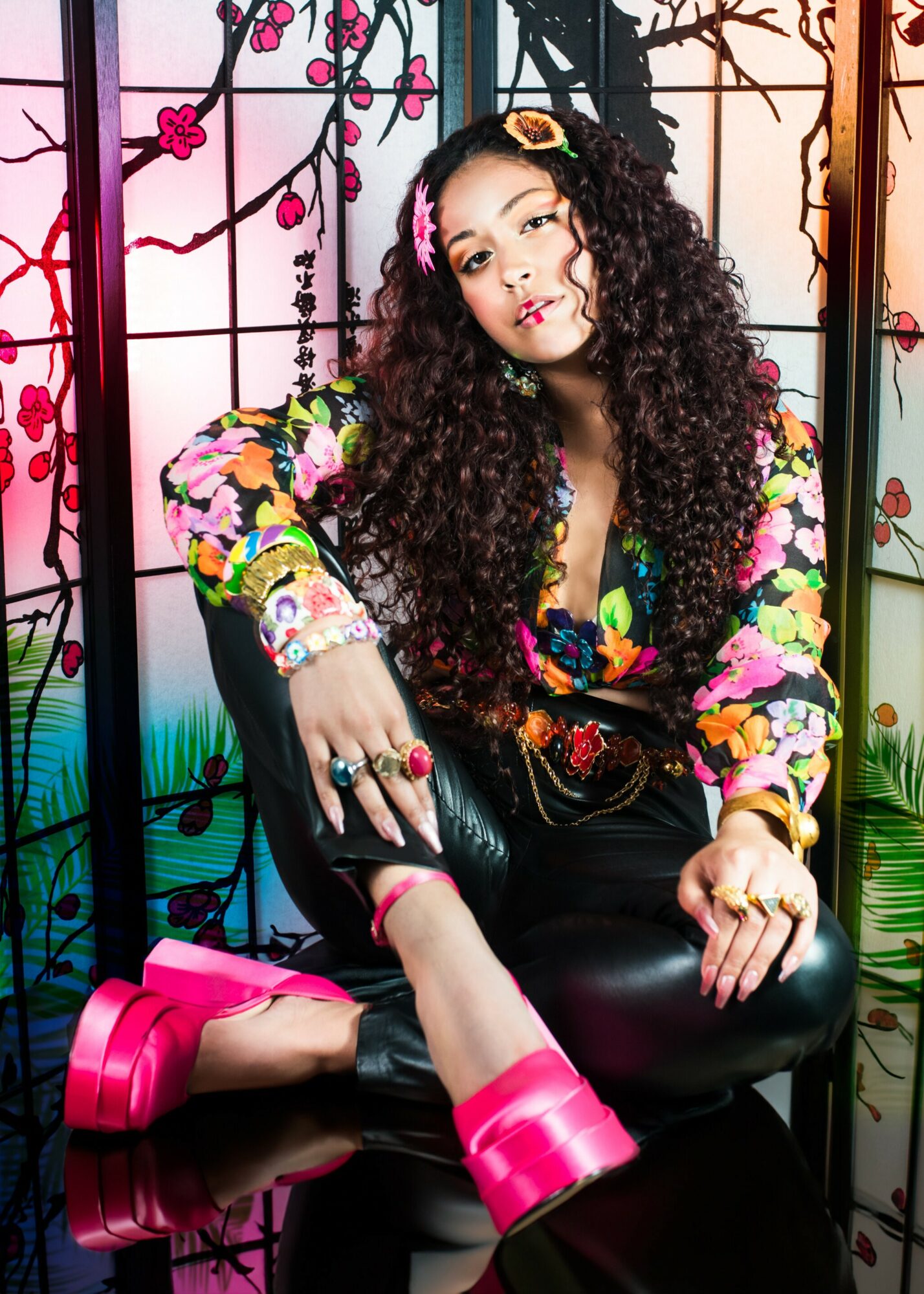 Young woman with curly hair, colorful floral jacket, sitting on a black chair, pink shoes, decorative background, holding a black bag.