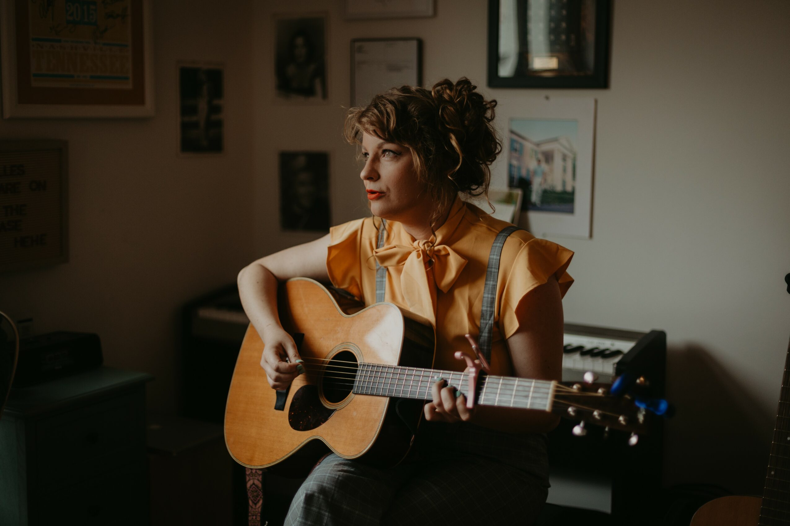 Woman with curly hair playing an acoustic guitar indoors, surrounded by framed pictures on the wall.