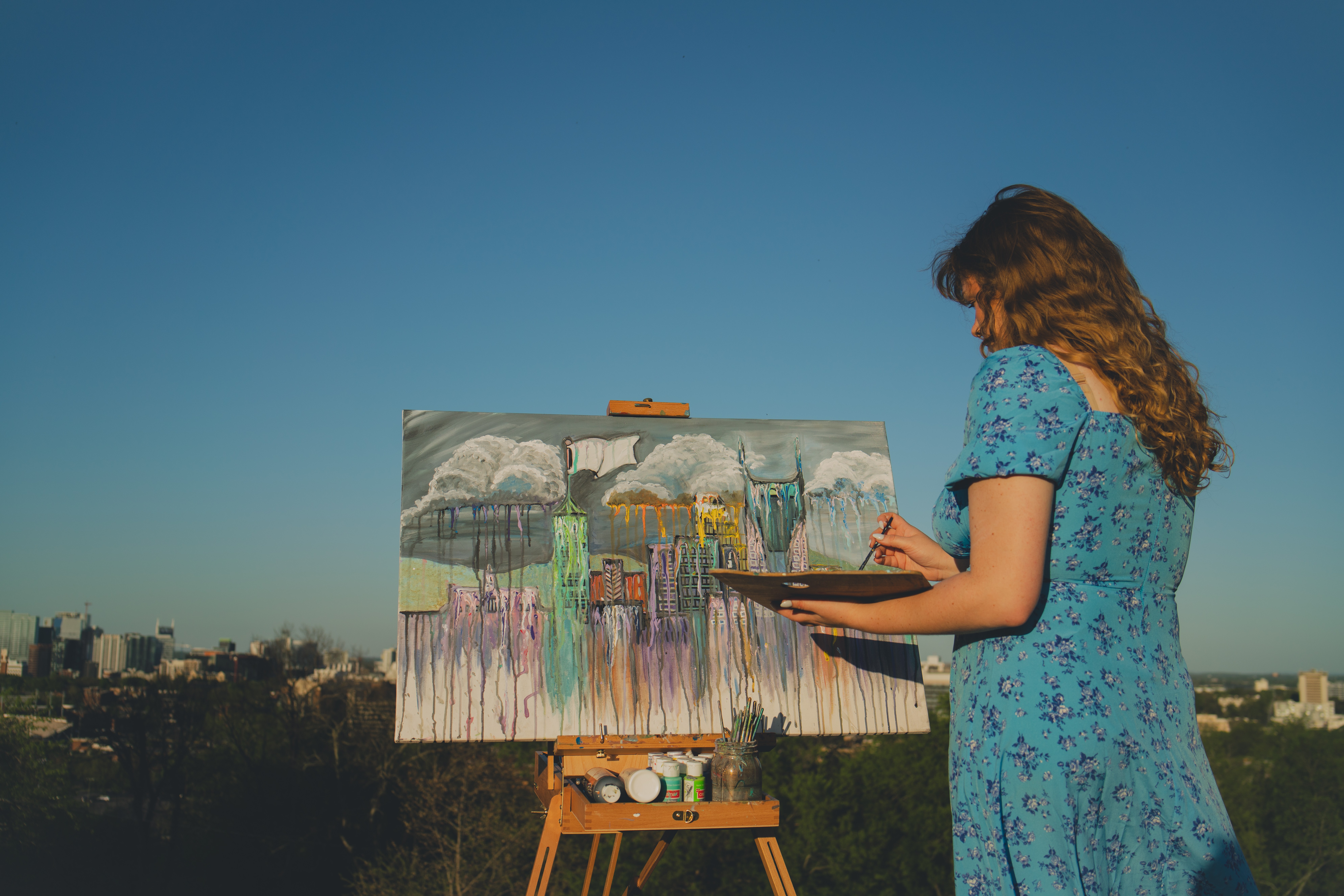 Woman with long curly hair in a blue floral dress paints on an outdoor canvas against a clear sky, cityscape in background.