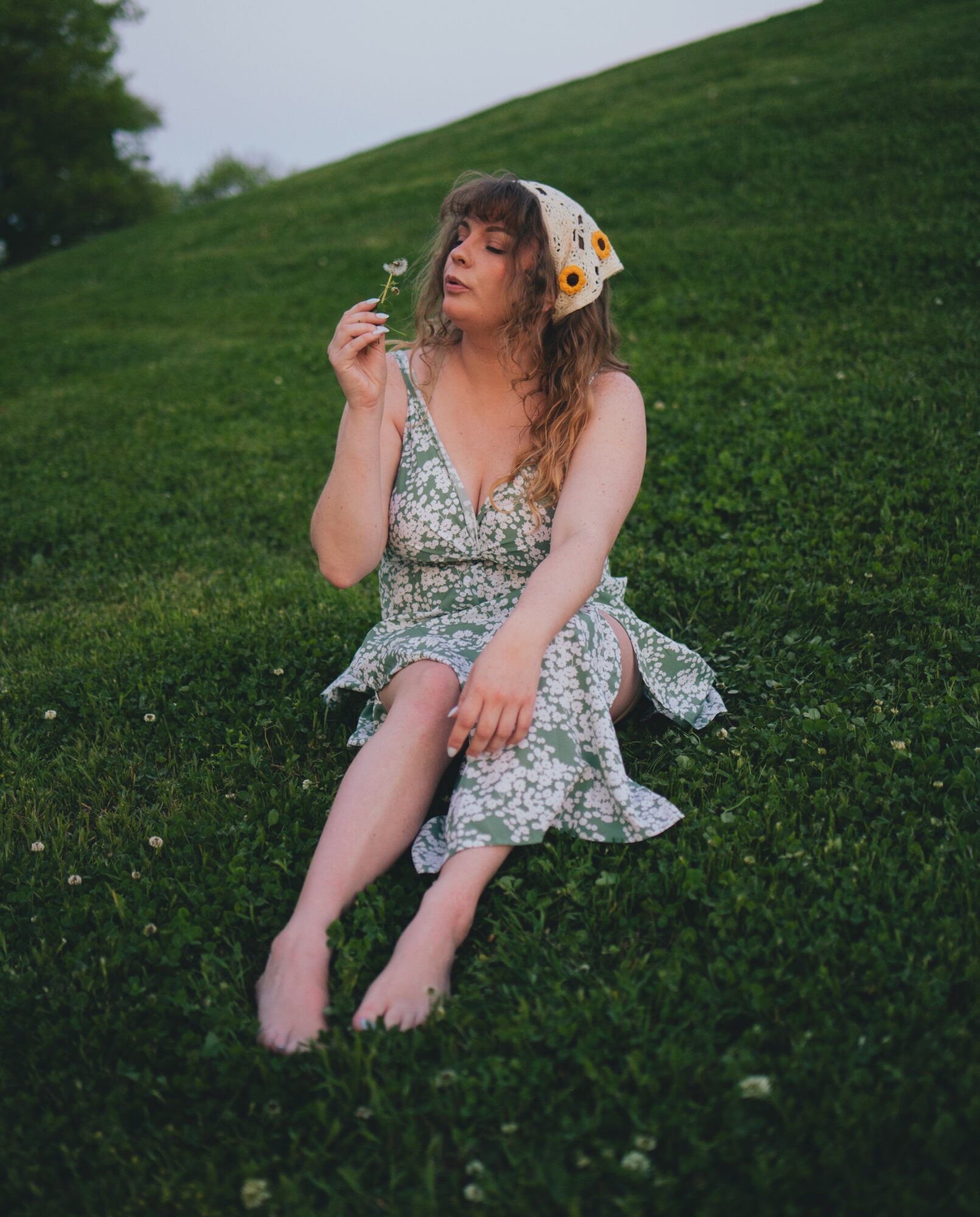 Young woman sitting on grass, holding a flower near her face, wearing a floral dress and a white headscarf with yellow dots.