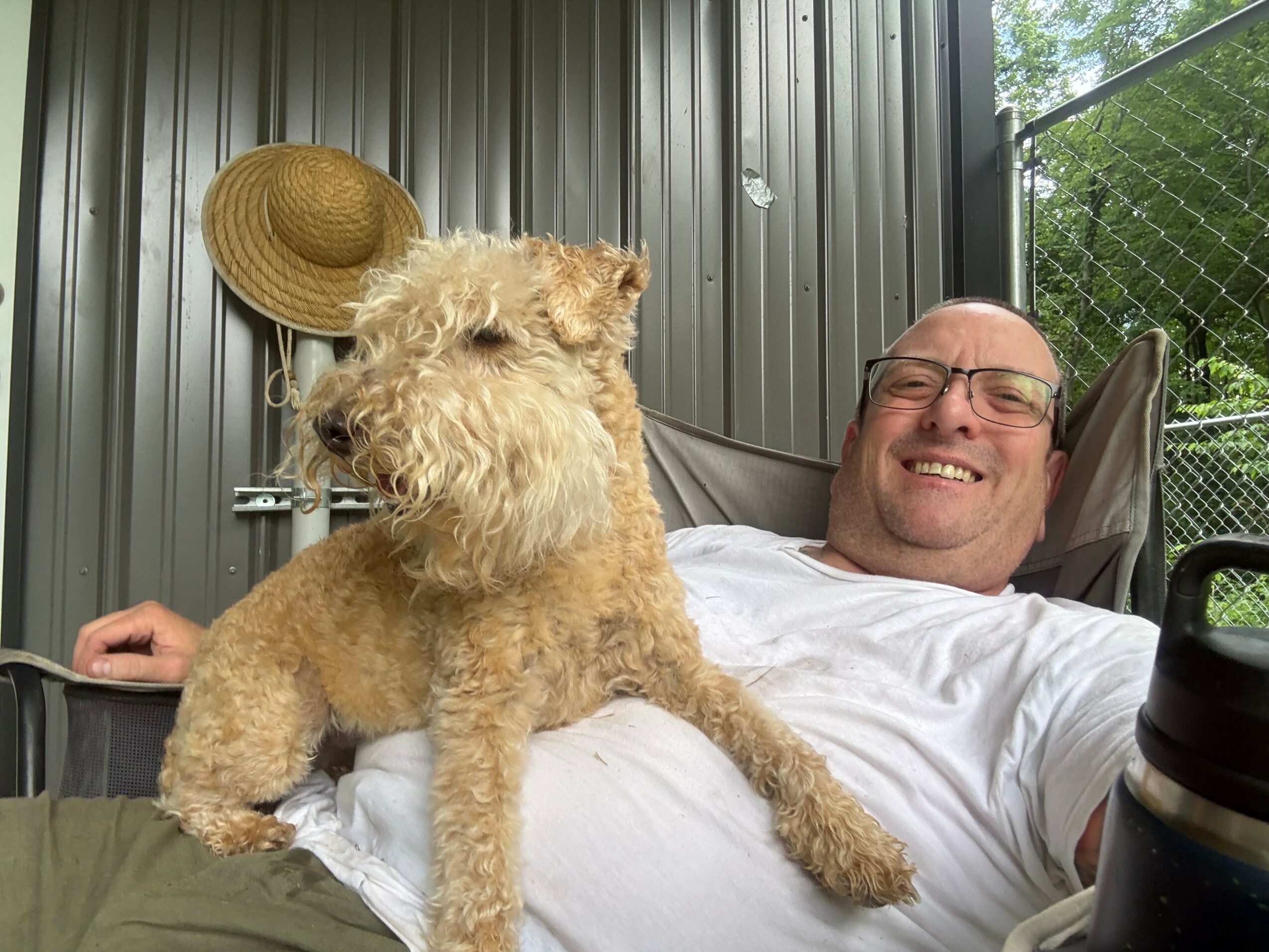 Man lying on a bed with a dog, both smiling, in a room with metal walls and a hat hanging on the wall.
