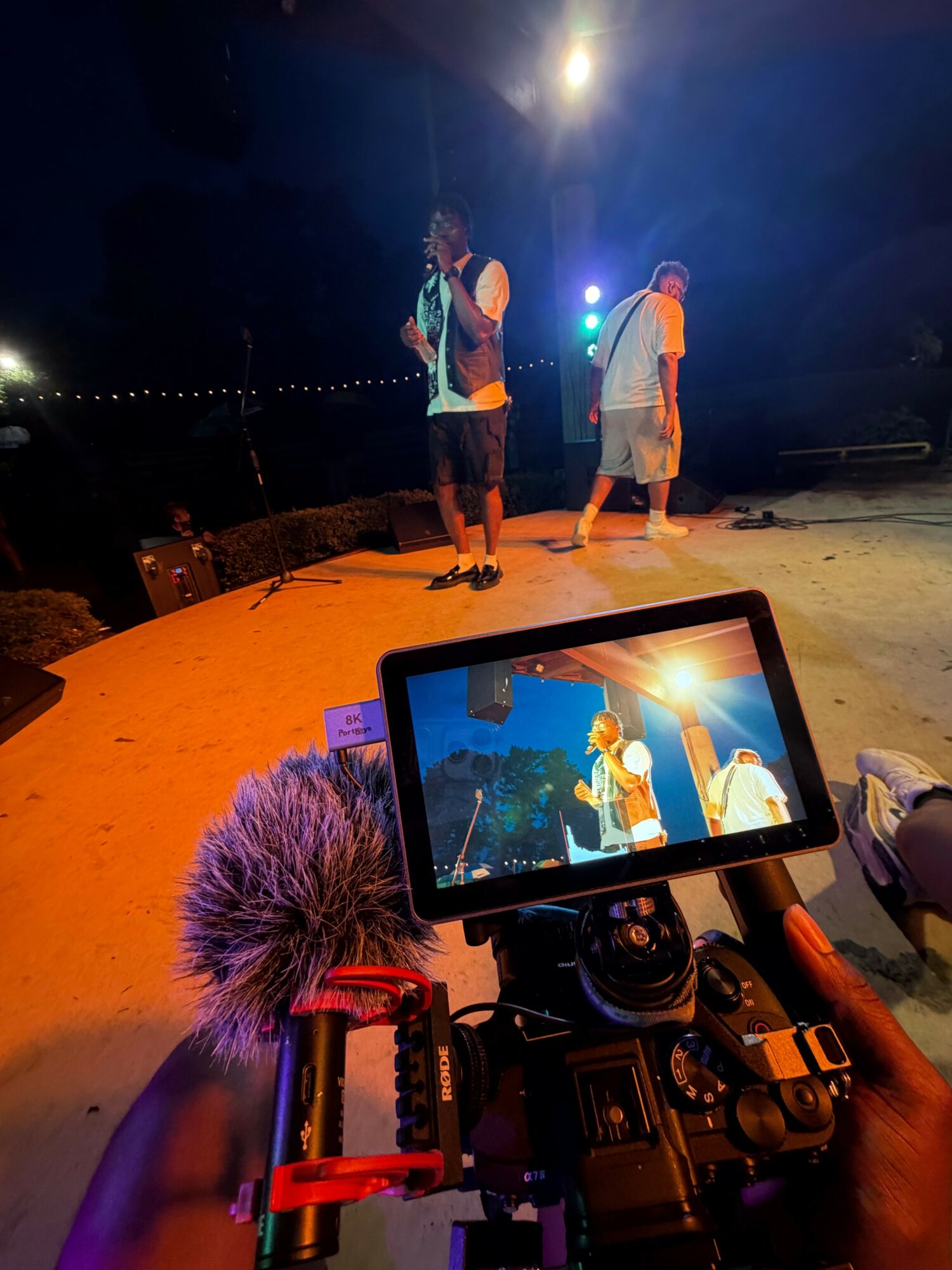 Three people stand outdoors at night, illuminated by a camera setup with a monitor showing a sunset scene.